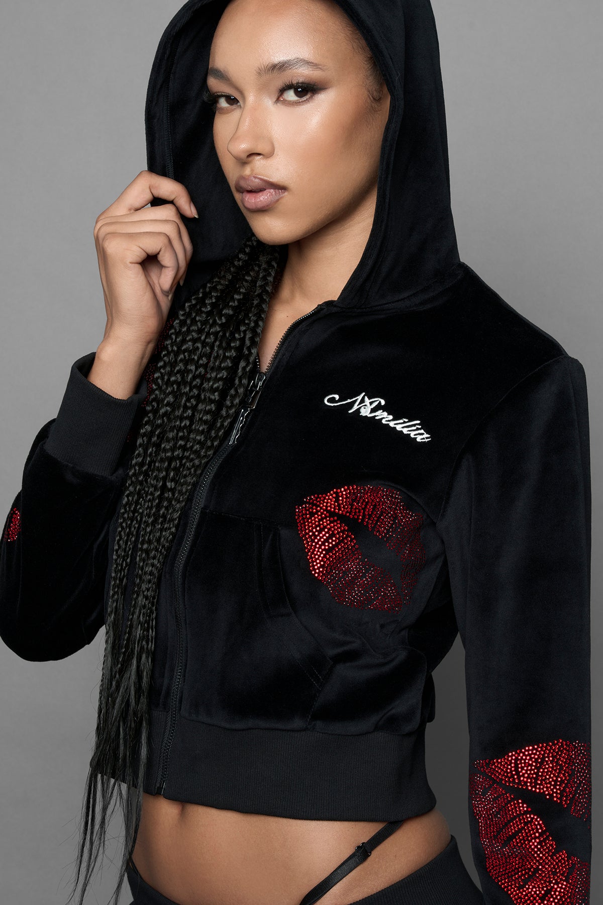 A female model wearing a black velvet zip-up hoodie with red sequined lip-print patches on the chest and sleeve, featuring white 'Namilia' embroidery on the left chest, photographed against a solid gray studio background.