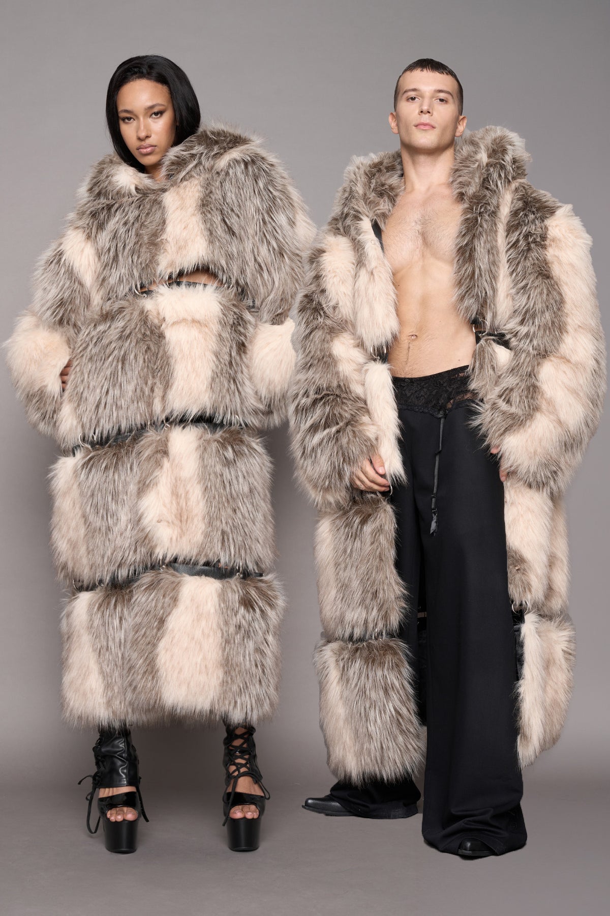 A man and a woman modeling matching oversized shaggy faux-fur coats in a studio setting. The coats are primarily light beige, cream, and gray with darker gray trim.