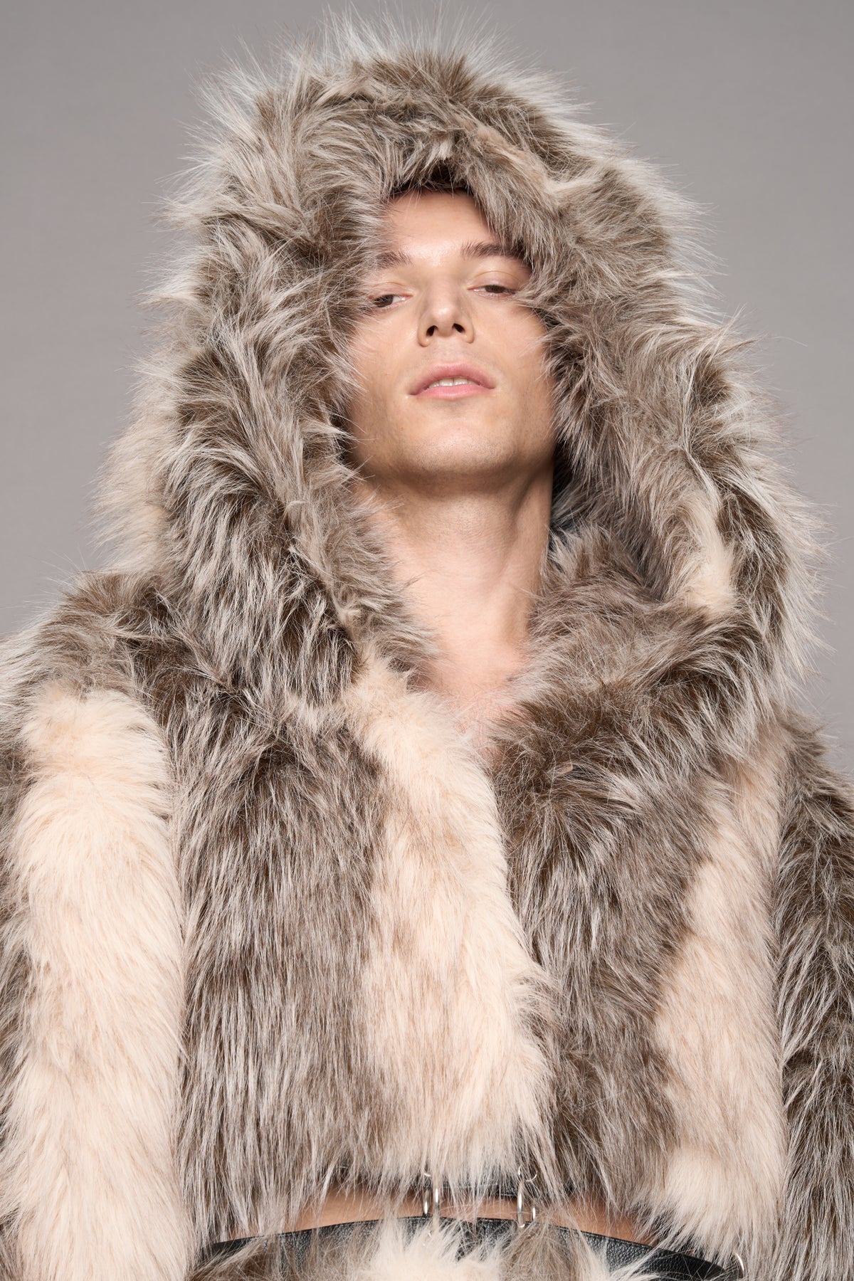 A close-up of a model wearing a luxurious hooded faux fur coat in mixed beige and gray tones, set against a solid gray studio background.