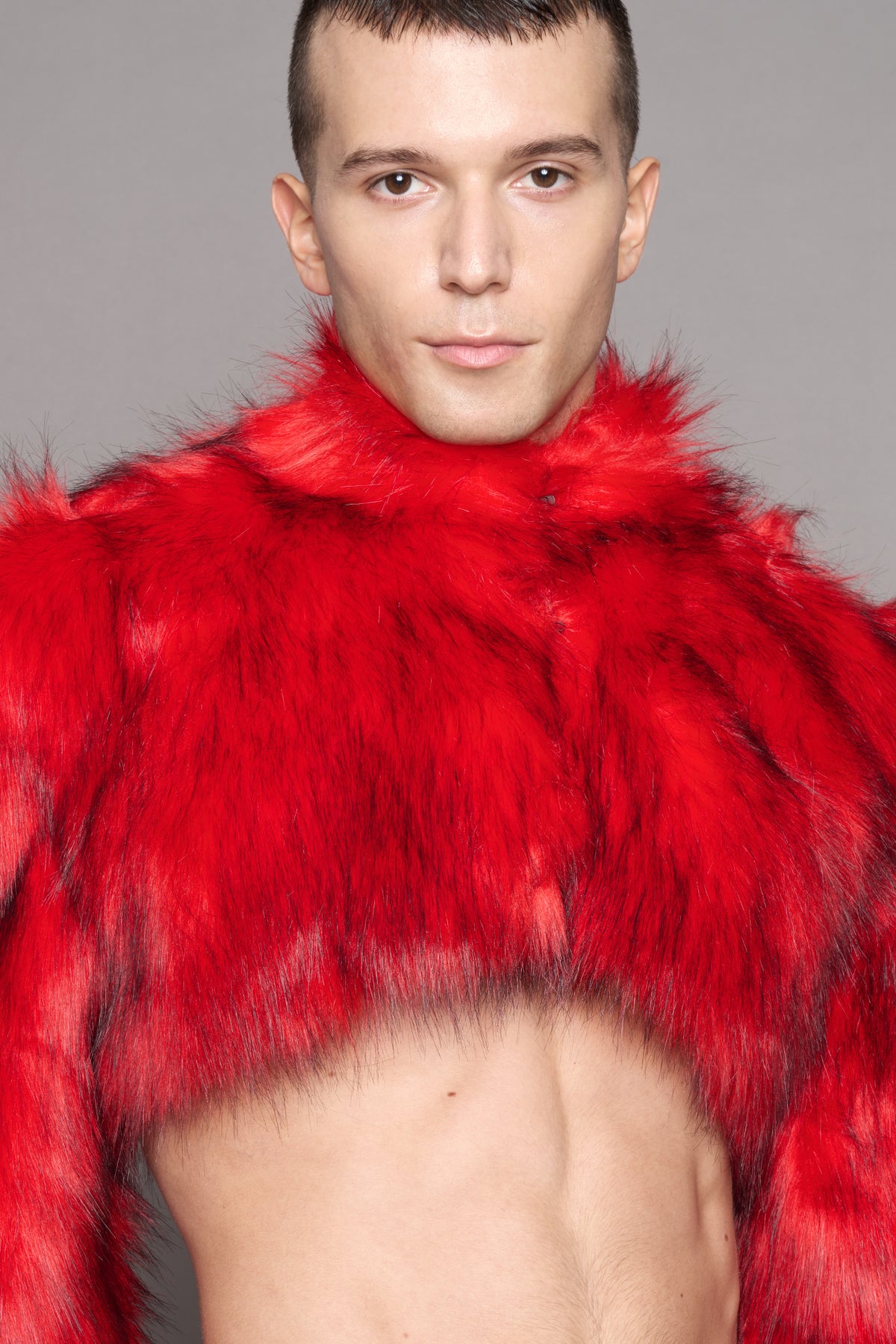 A close-up portrait of a man wearing a bright red faux-fur cropped jacket against a plain gray studio background.