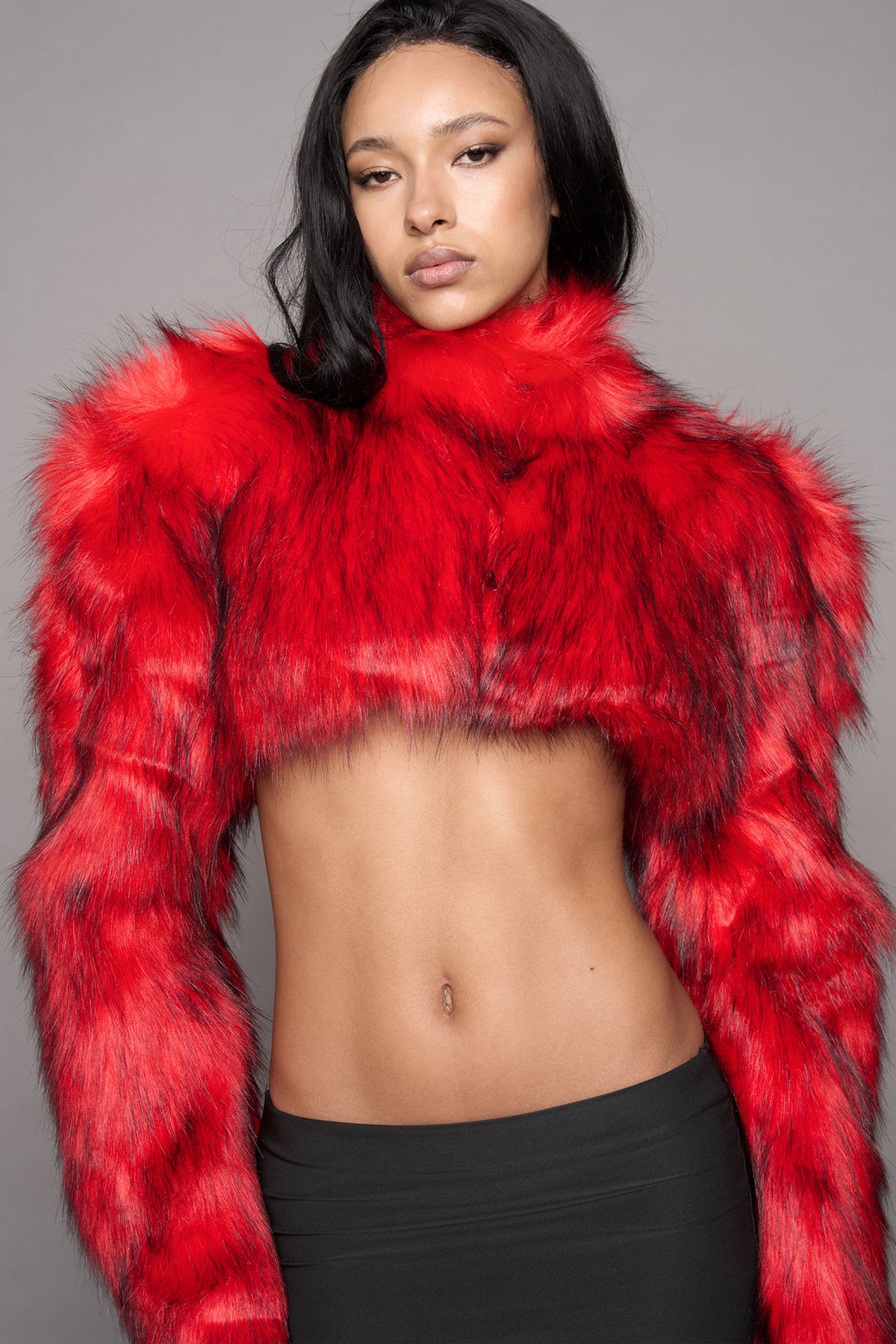 A close up of a model wearing a vibrant red faux fur cropped long-sleeve top with a high collar, paired with a black skirt, against a solid gray studio background.