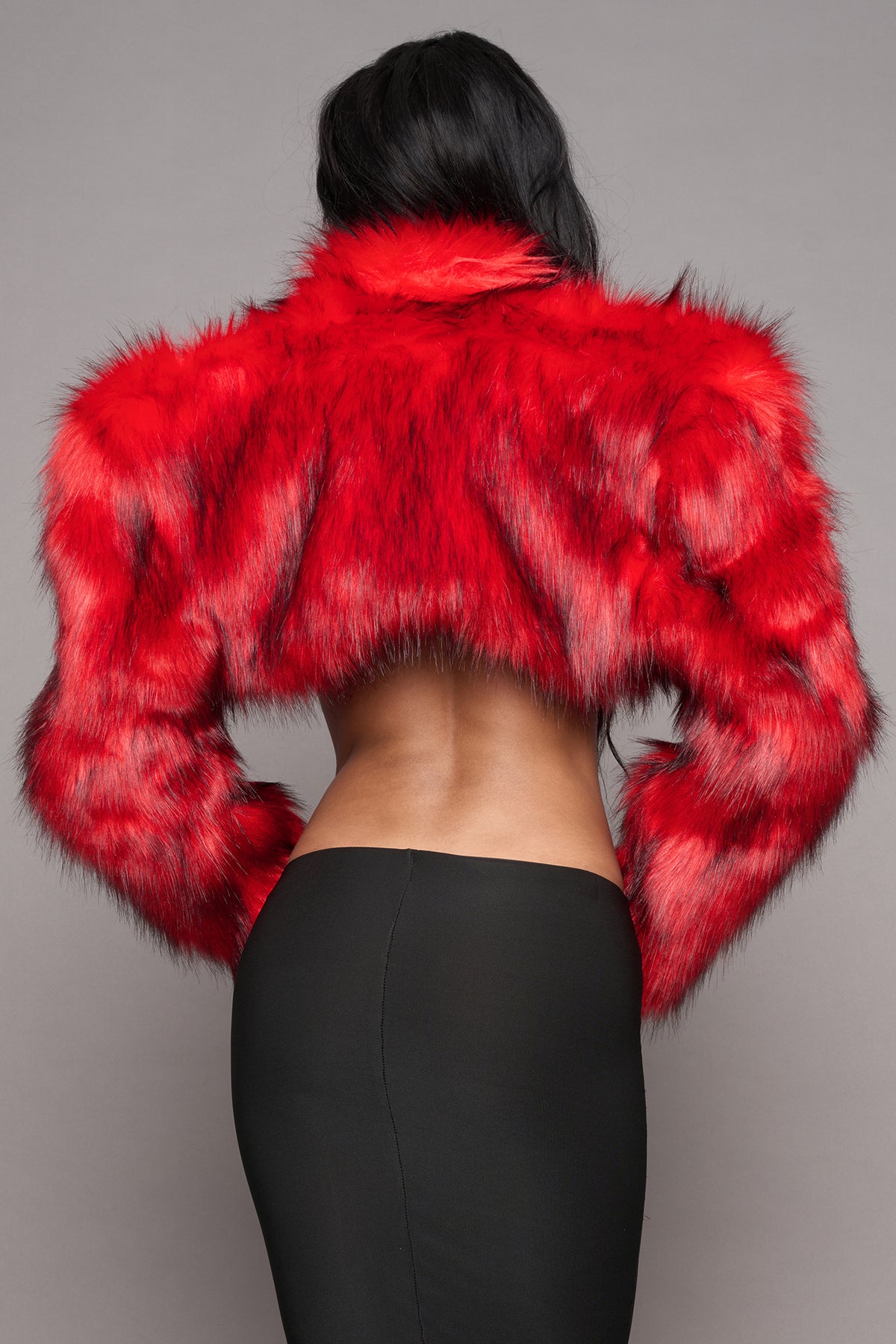 A model shown from the back wearing a vibrant red cropped faux fur jacket and black skirt, set against a solid gray studio background.