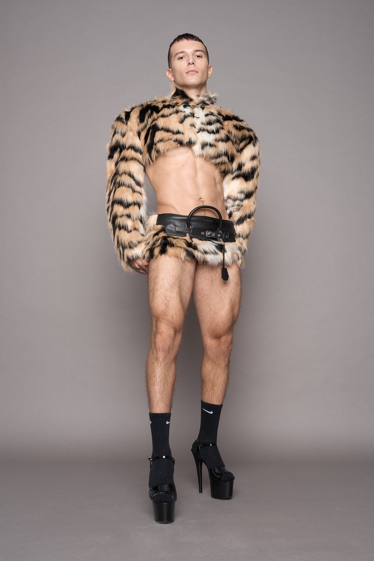 A model stands against a solid gray studio background wearing a cropped black and tiger faux fur jacket, a tiger faux fur mini skirt, black socks, and black platform heels.