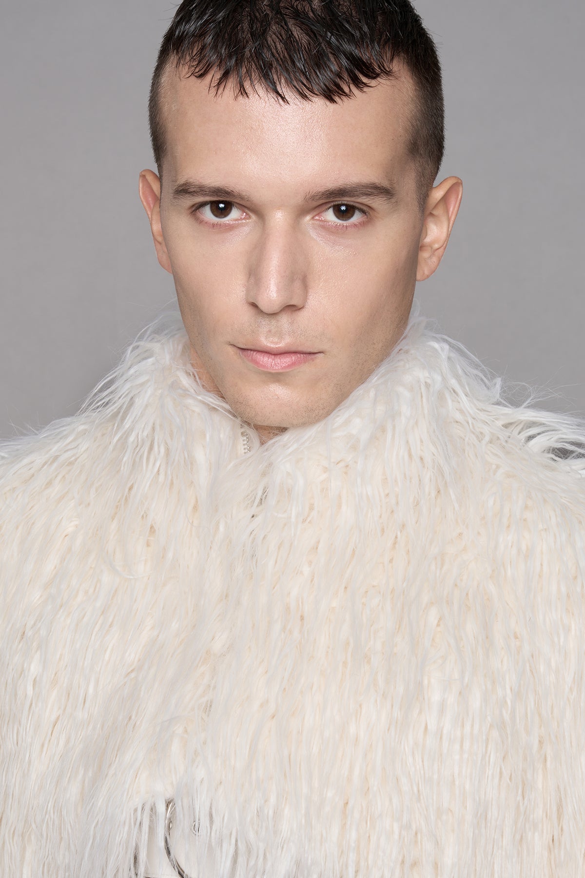 A close-up portrait of a man wearing a voluminous, shaggy white faux fur collar or jacket against a plain gray studio background.
