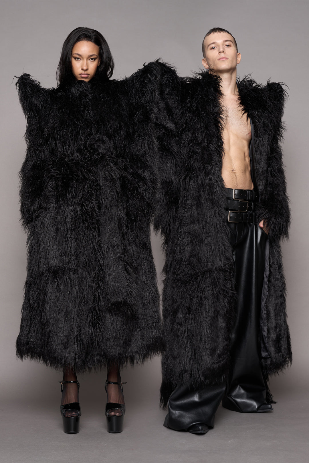 Two models wearing oversized black shaggy faux-fur coats with spiky constructed shoulders with a matching fur waist tie 
