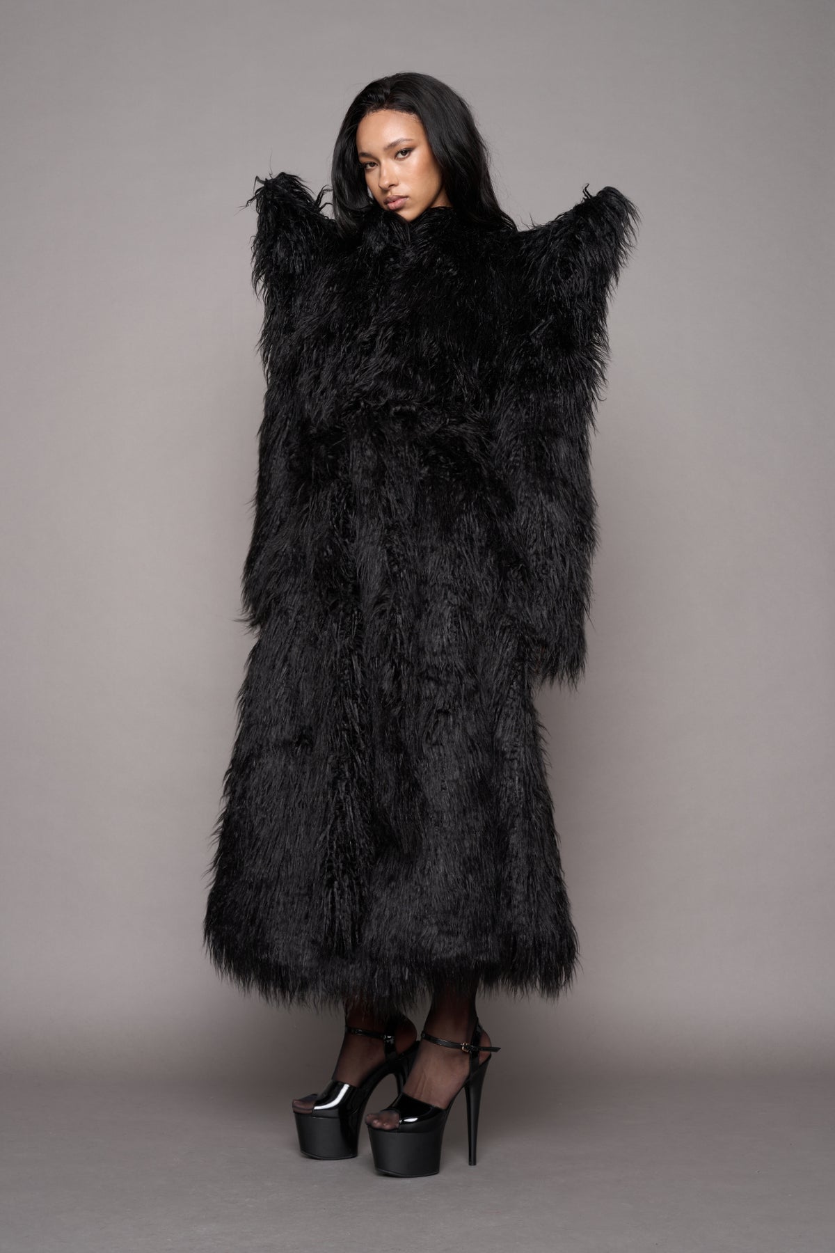 A female model wearing a voluminous black shaggy faux-fur coat with dramatic shoulder spikes closed with a matching waist tie, styled with black platform heels against a neutral gray studio background.