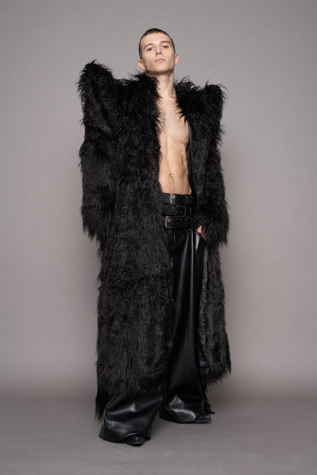 A model stands against a plain gray studio background wearing a voluminous black faux fur coat over a shirtless and black wide-leg leather pants.