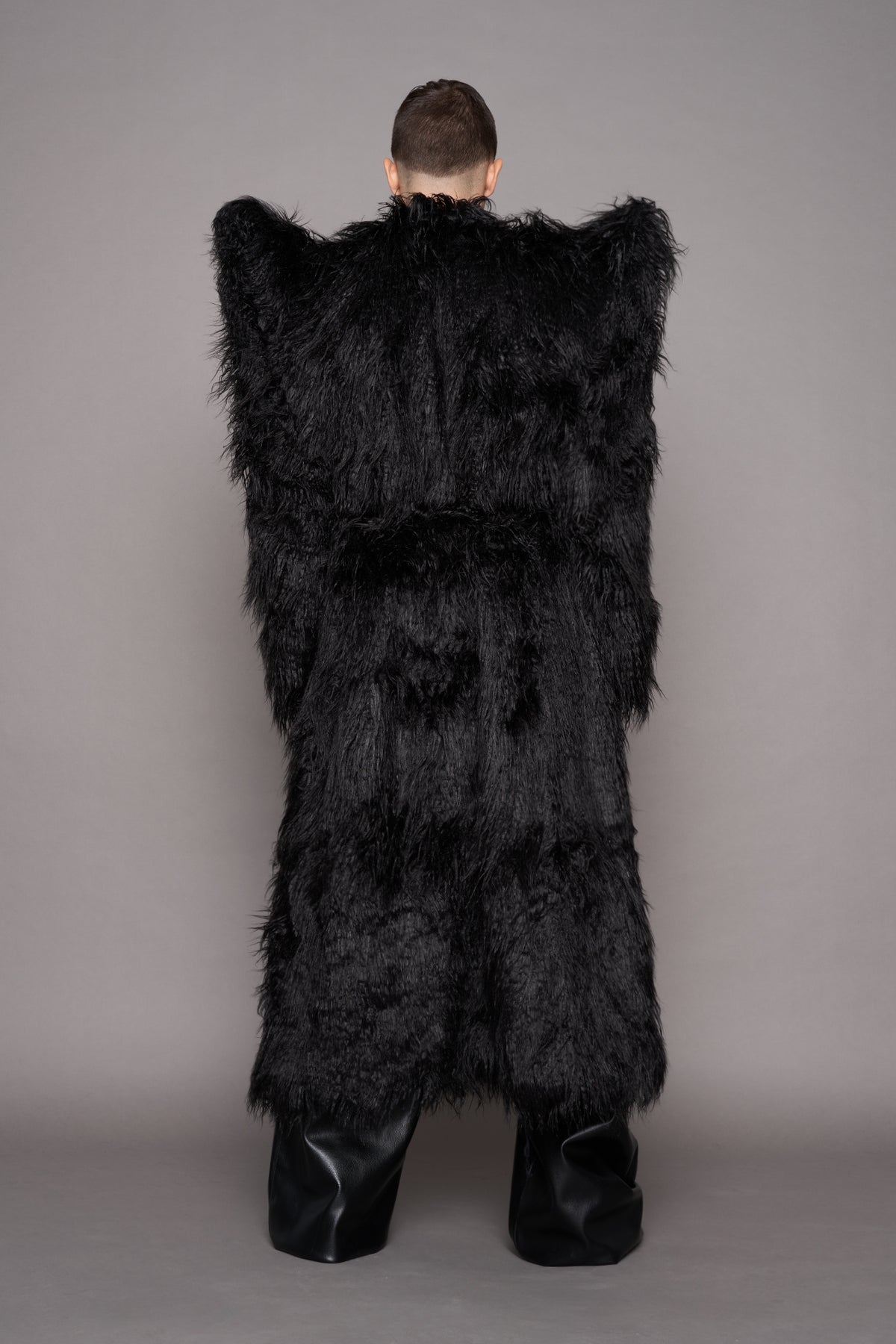 A model viewed from the back wearing a voluminous black shaggy faux-fur coat with wide shoulder panels and black boots, standing against a plain gray studio background.