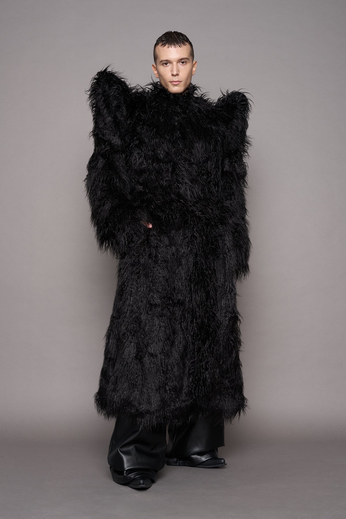 A model stands against a plain gray studio background wearing a voluminous black shaggy faux-fur coat with dramatic spiky shoulder and black wide-leg pants.