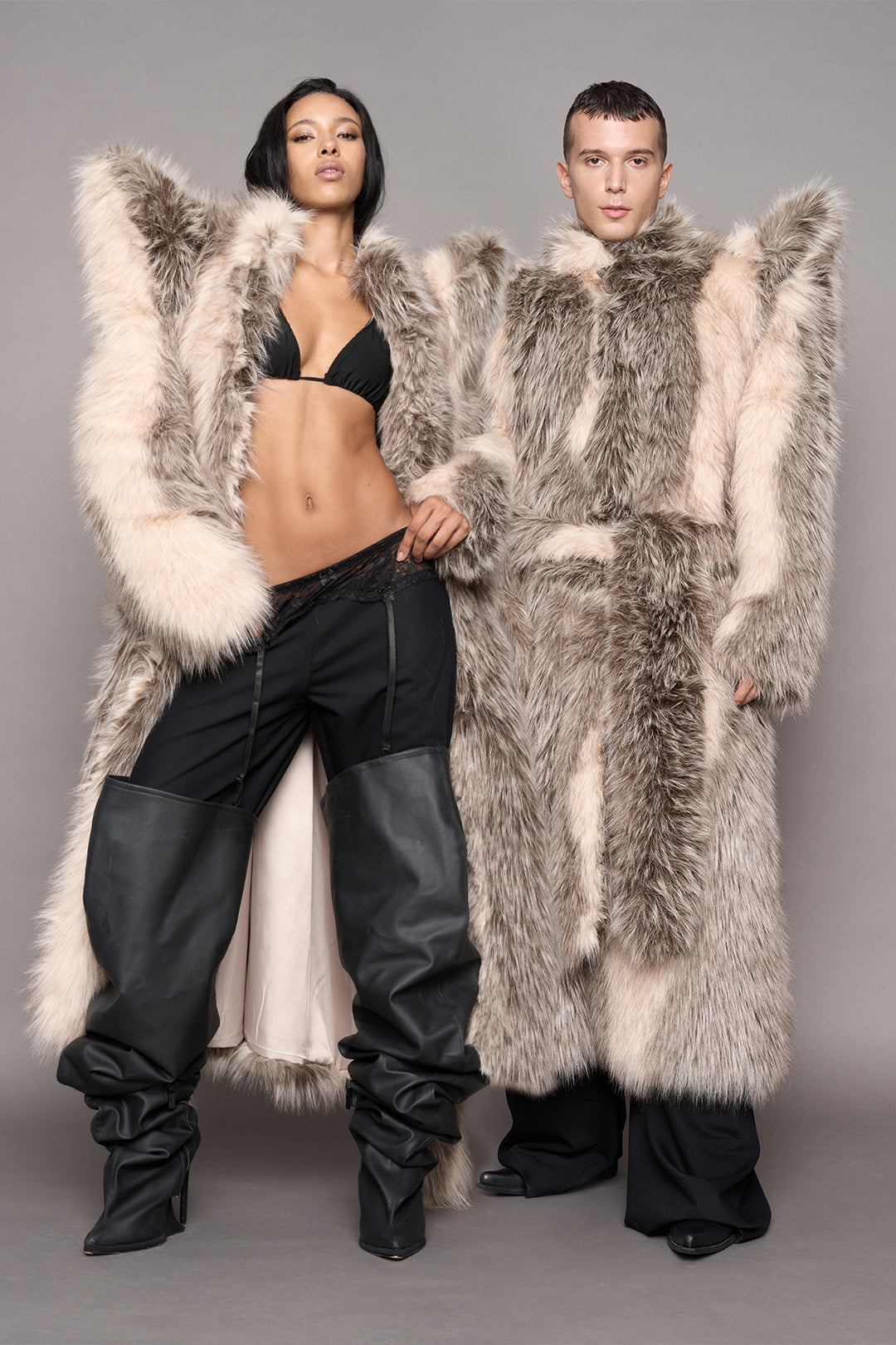 Two models wearing oversized, dramatic faux-fur coat in gray, beige, and cream tones, styled with black pants and black leather boots against a plain gray studio background.