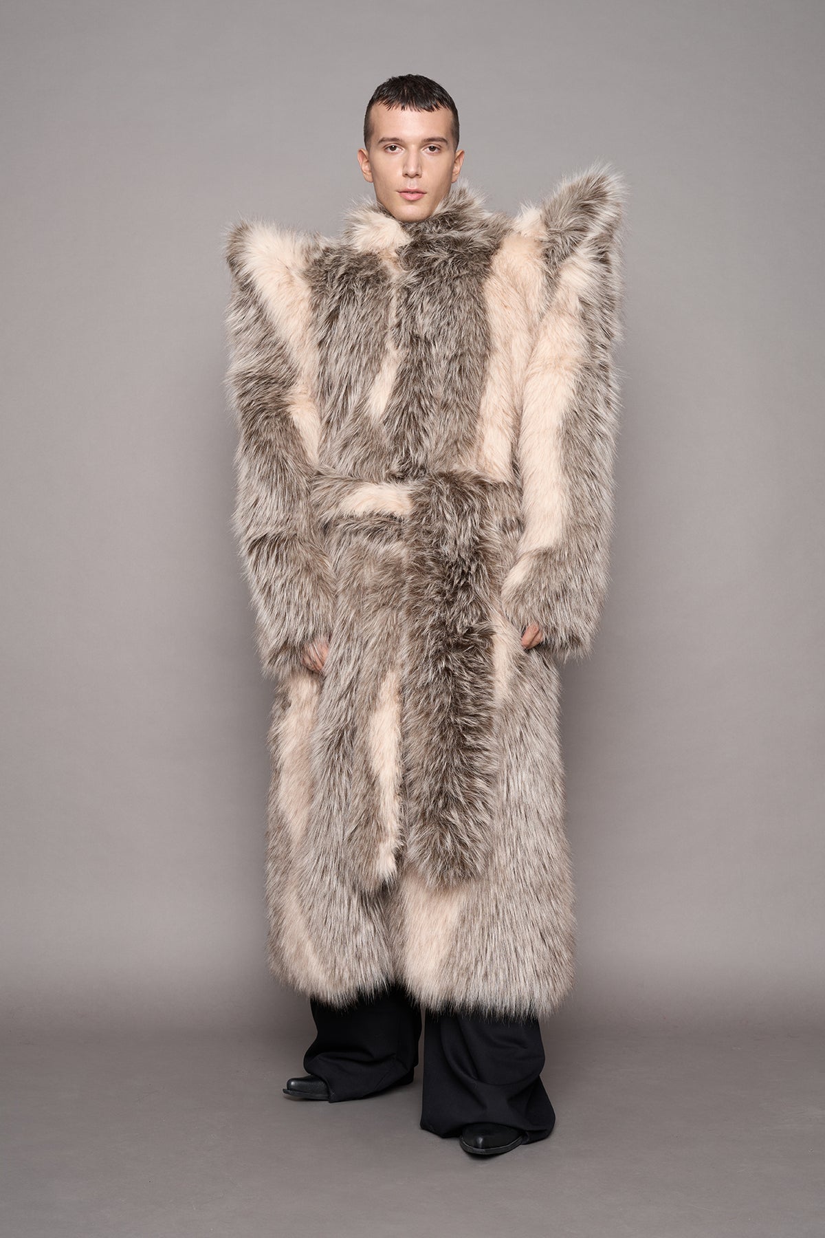 A model poses in a closed long, oversized black faux-fur coat with spikey shoulders, black pants and black boots, set against a solid gray studio background.