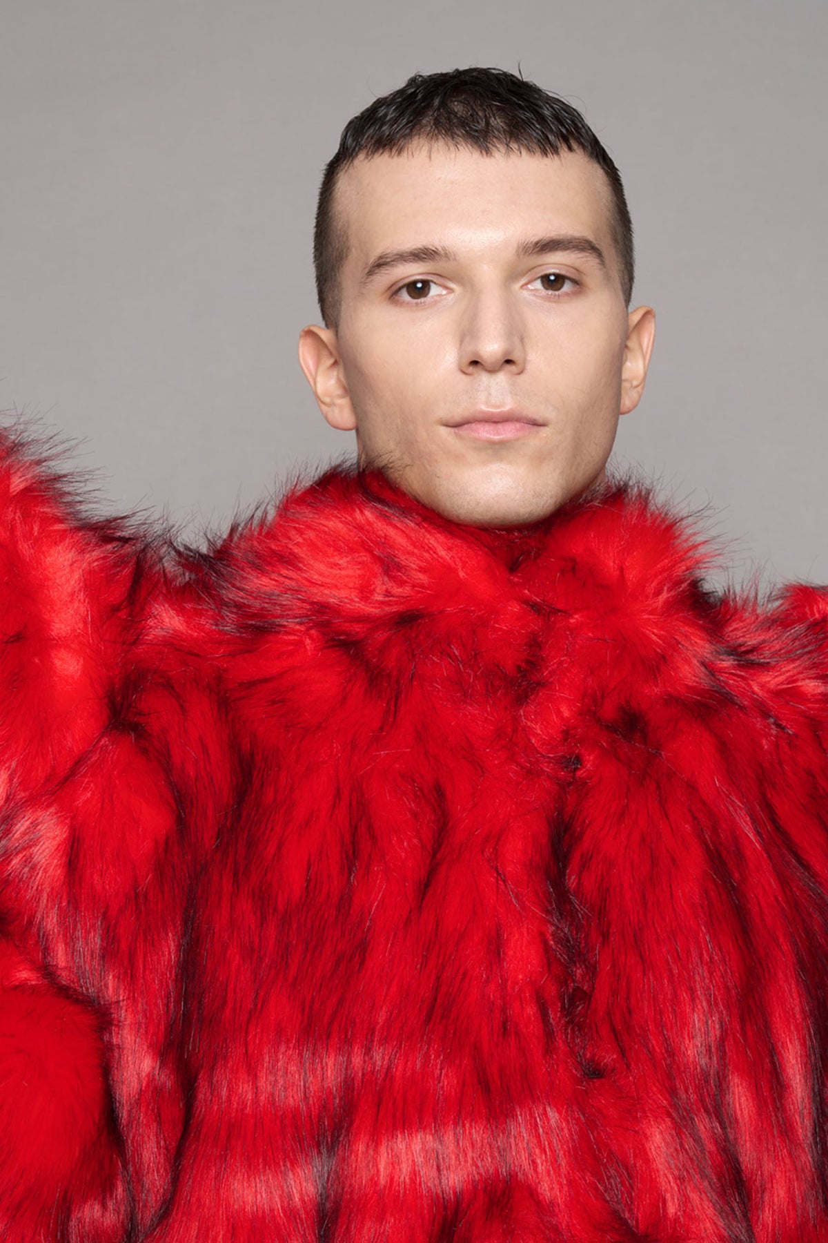 A close-up portrait of a man wearing a voluminous, bright red fur coat against a solid gray studio background.