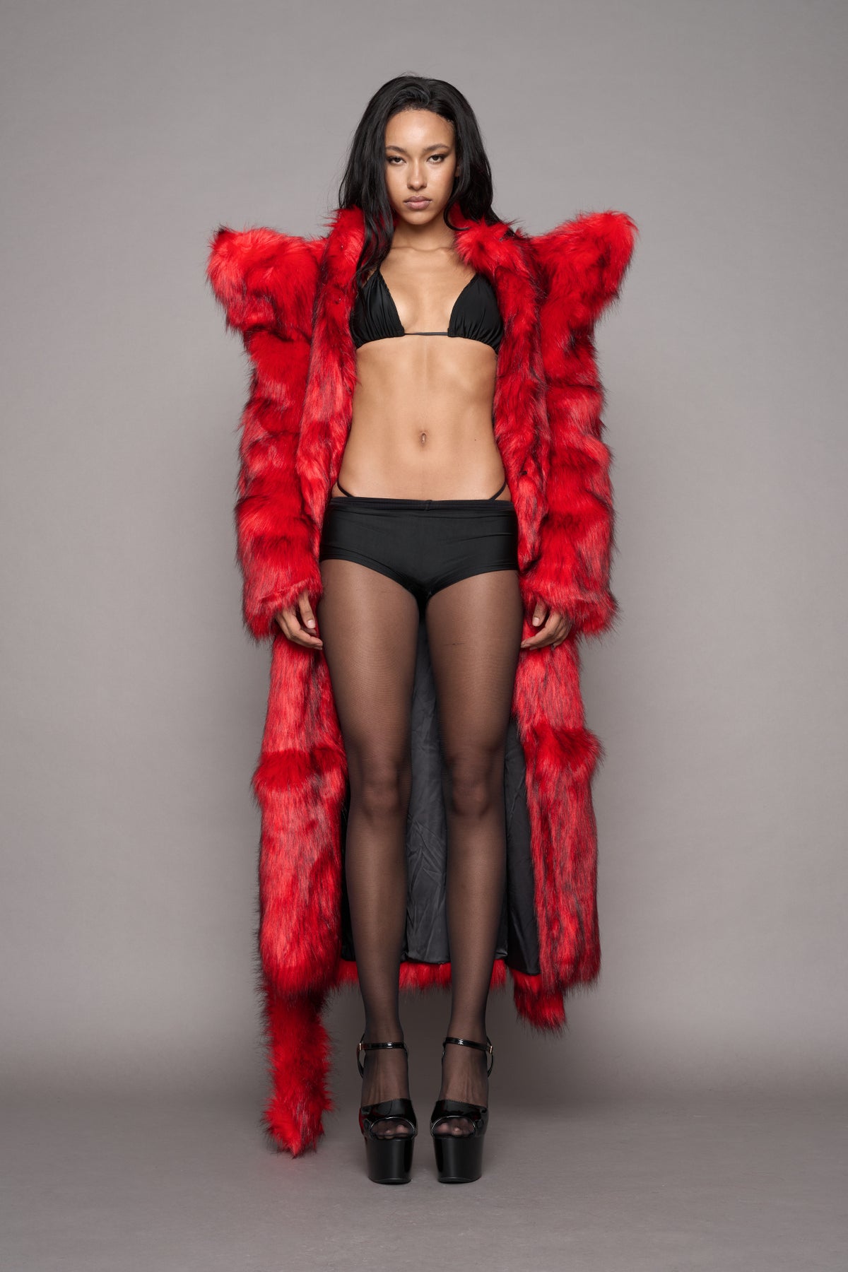 A model stands against a plain gray studio background wearing a bright red shaggy faux-fur coat with big spiky shoulders and a hanging matching waist tie over a black bikini, black sheer tights, and black platform heels.