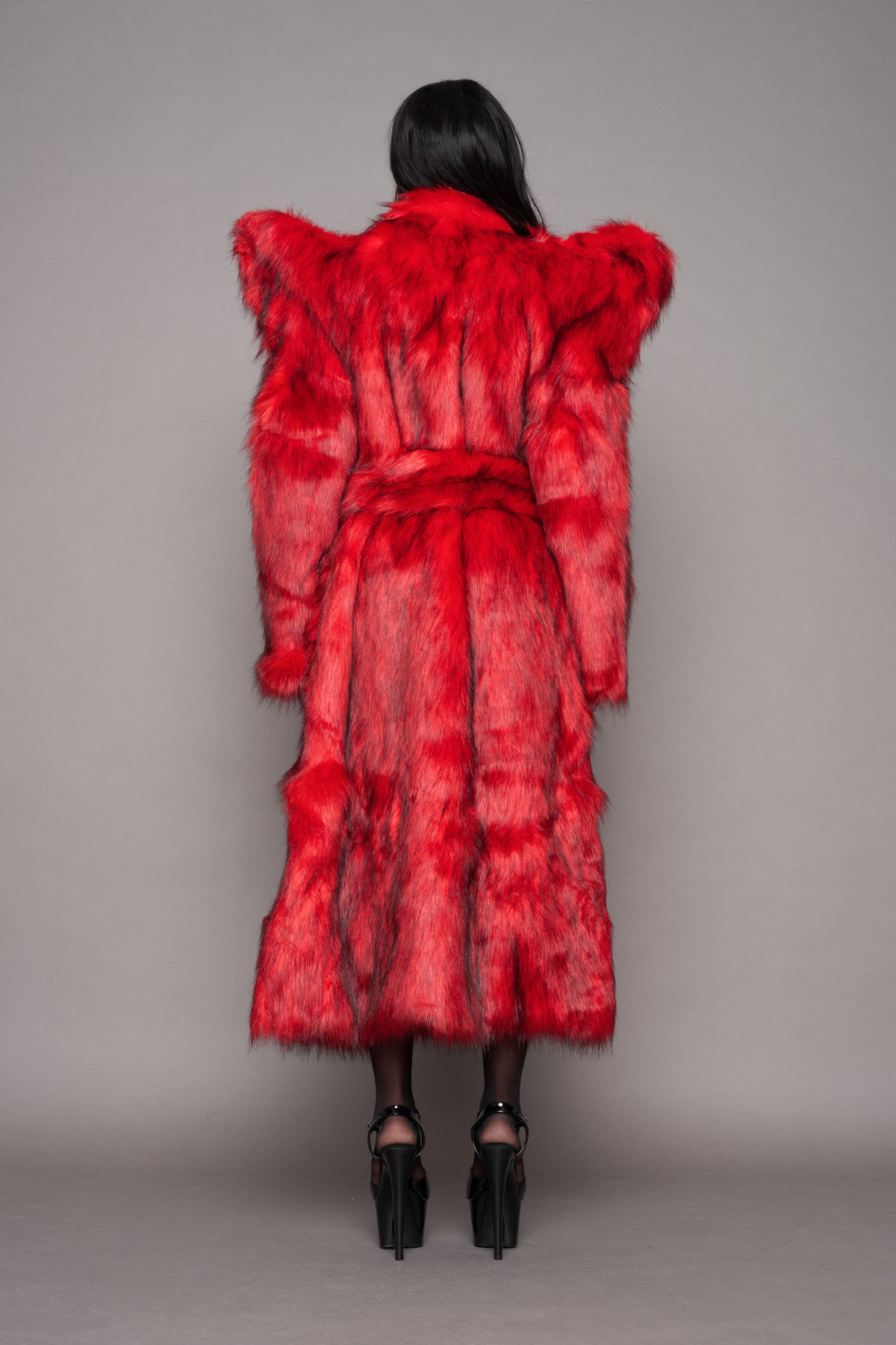 A model viewed from behind wearing a voluminous, bright red faux fur coat with dramatic shoulder spikes and a matching waist tie, paired with black high heels against a neutral gray studio background.