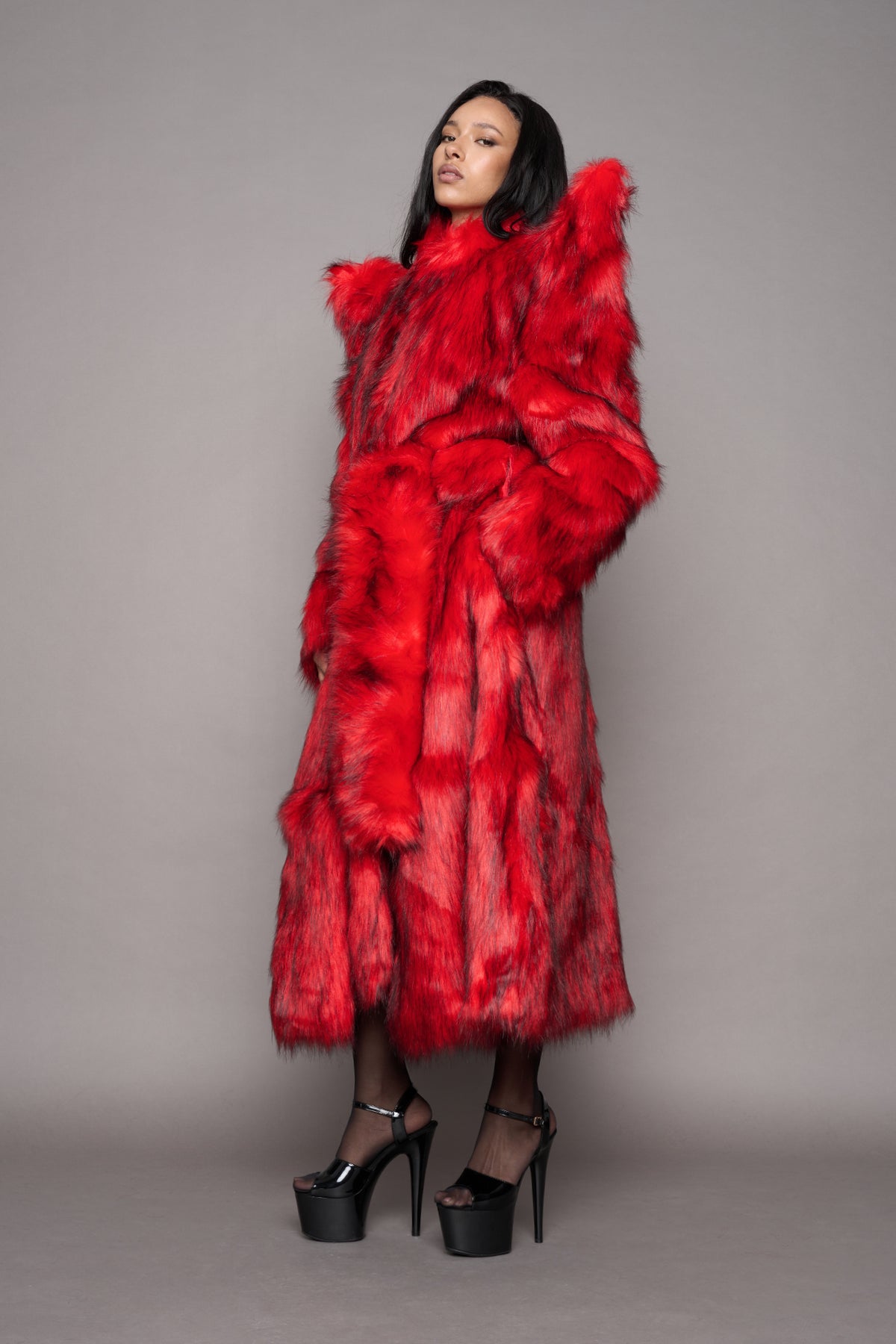 A model wearing a voluminous, bright red fur coat with dramatic shoulder spikes and a matching waist tiepaired with black high-heeled platform sandals, set against a solid gray studio background.