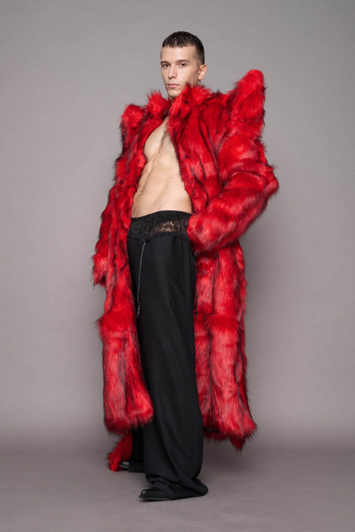 A male model wearing a dramatic, oversized bright red faux fur coat with dramatic shoulder spikes, worn open over a black lace-trimmed black pants standing against a solid gray studio background.