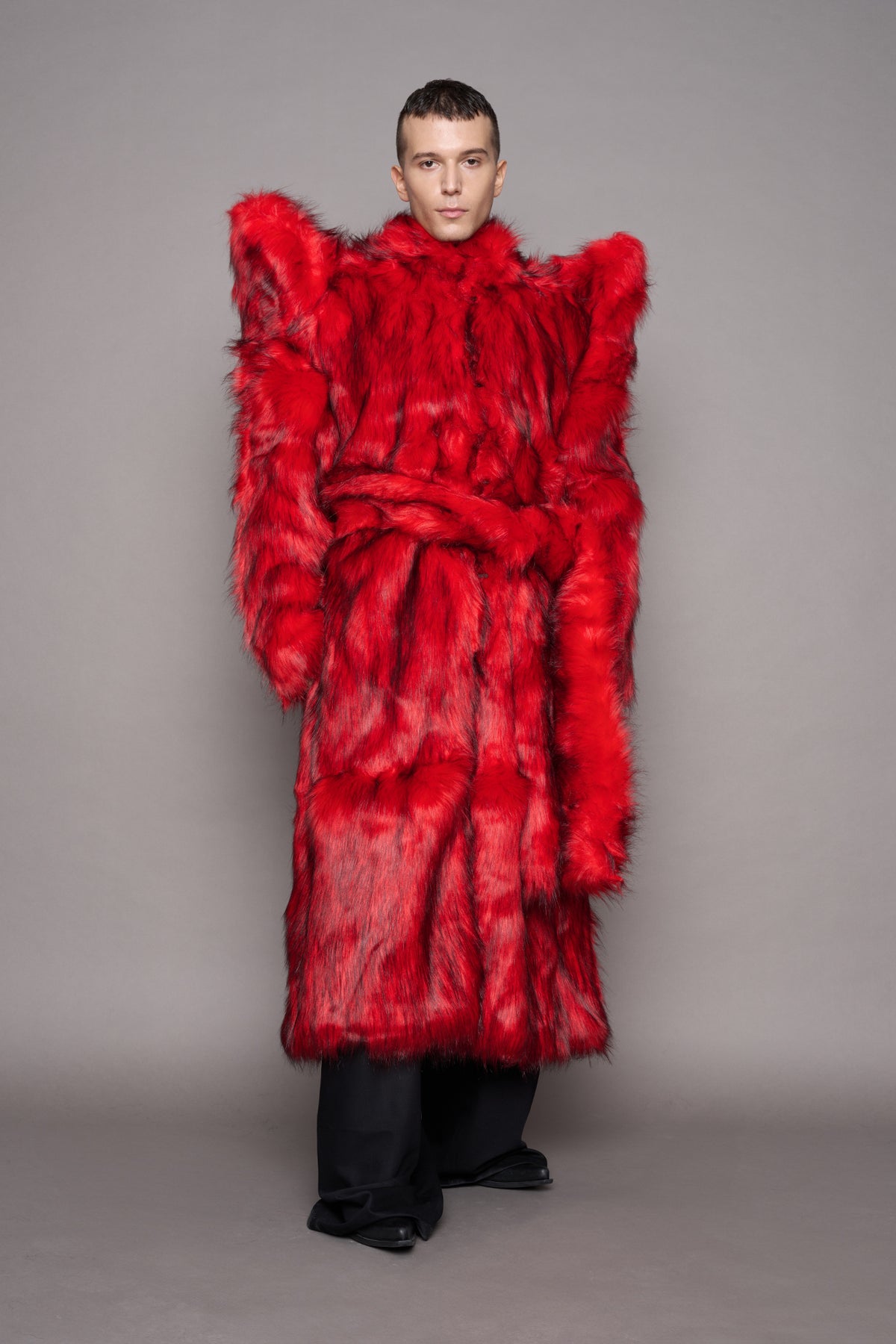 A model stands against a plain gray studio background wearing an oversized, voluminous bright red faux-fur coat with dramatic shoulder spikes and a matching belt, paired with loose black pants and black shoes.