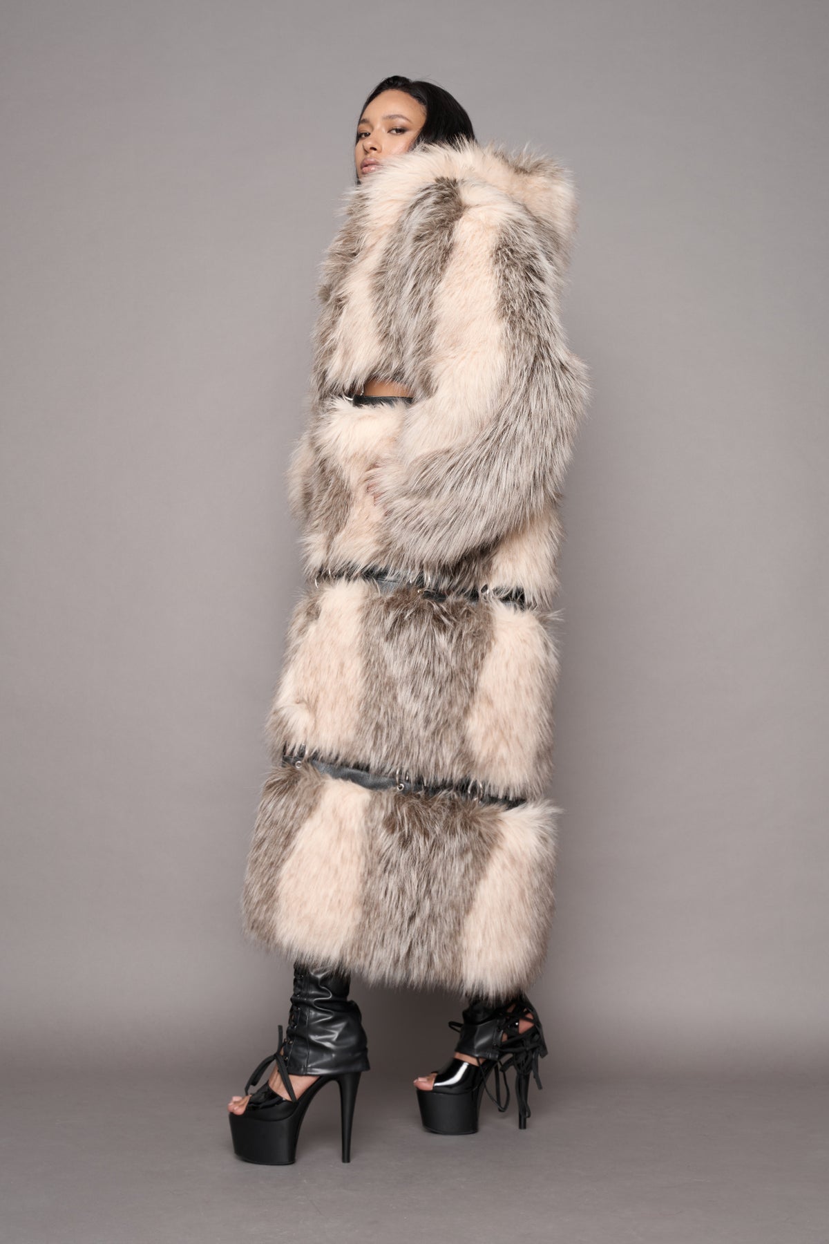 A model wearing a long faux fur coat with black leather trim and black high-heeled platform sandals, photographed against a solid gray studio background.