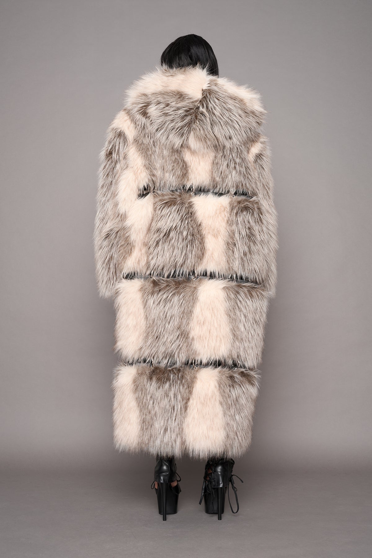 A model shown from the back wearing a long, oversized faux fur coat with a hood in shades of cream, beige, and gray, paired with black high-heeled boots against a solid gray studio background.