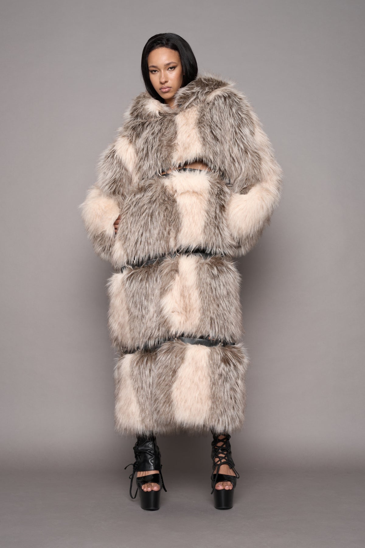 A model wearing a voluminous faux fur coat in shades of gray, cream, and white, paired with black platform lace-up heels, shot against a solid gray studio background.
