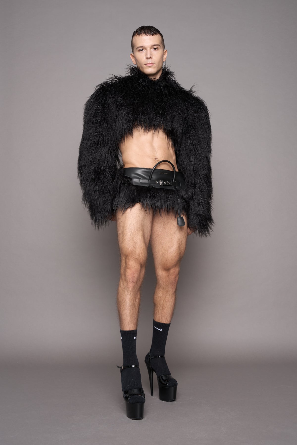 A male model stands against a plain gray studio background wearing a voluminous black furry jacket with a high collar, a black faux fur miniskirt with a handbag handle attached in the front , paired with black crew socks, and black platform high-heel shoes.