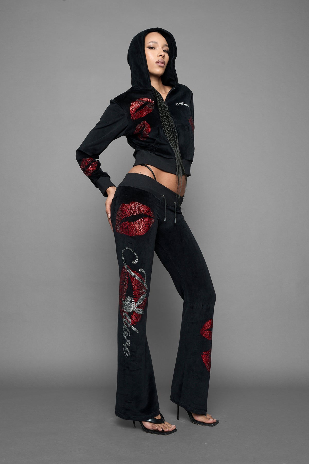 A female model wearing a black velvet zip-up hoodie with red sequined lip-print patches on the chest and sleeve, featuring white 'Namilia' embroidery on the left chest, photographed against a solid gray studio background.