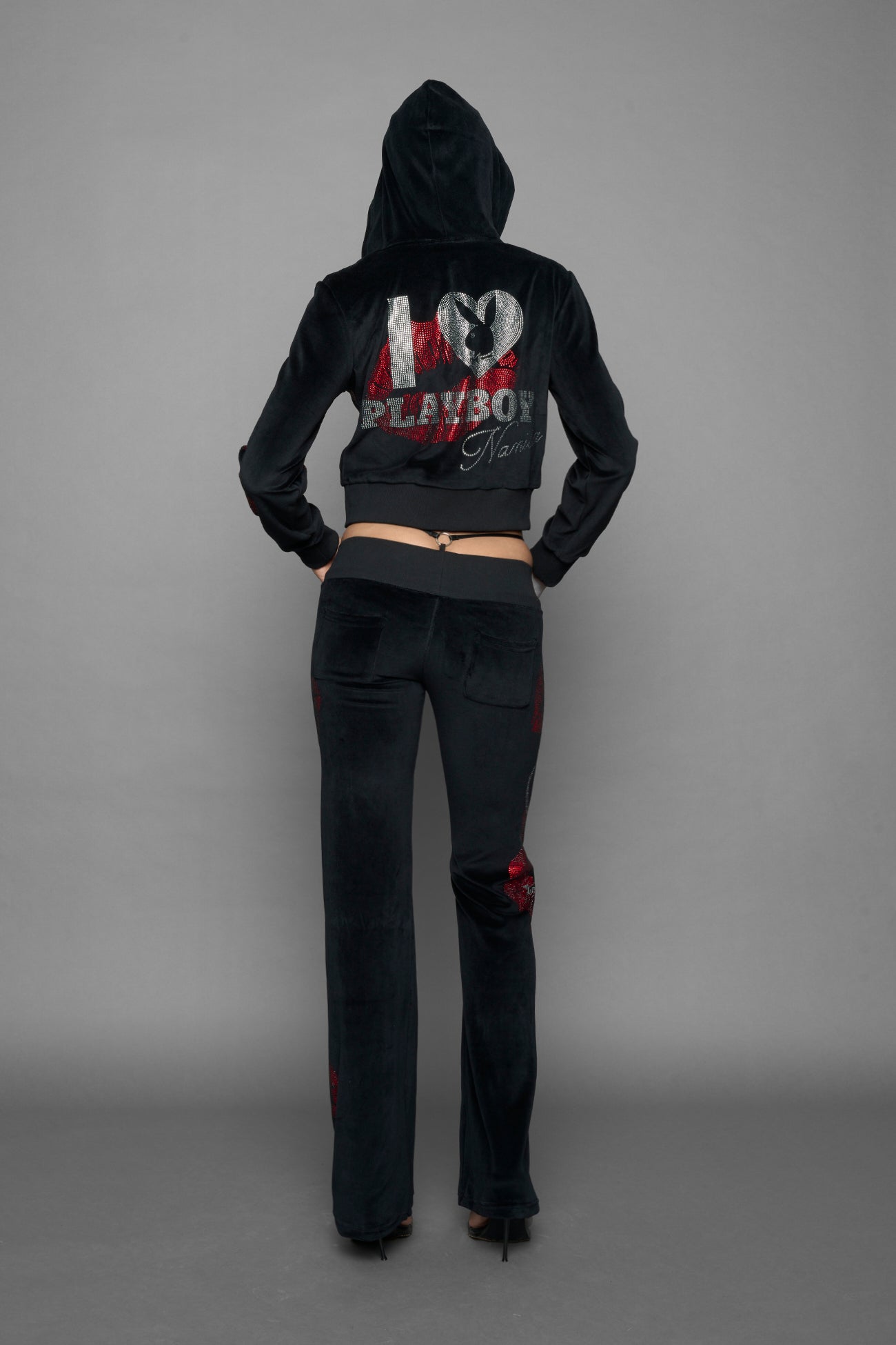 A female model wearing a black velvet zip-up hoodie with red sequined lip-print patches on the chest and sleeve, featuring white 'Namilia' embroidery on the left chest, photographed against a solid gray studio background.