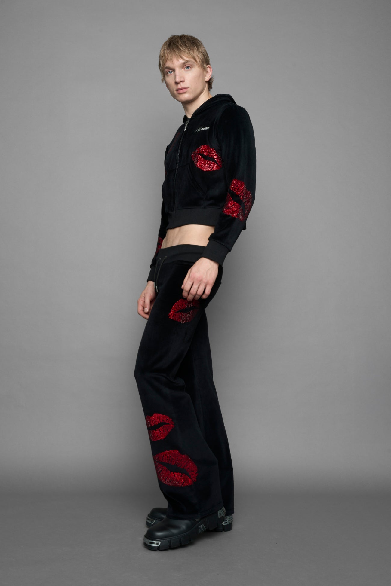 A female model wearing a black velvet zip-up hoodie with red sequined lip-print patches on the chest and sleeve, featuring white 'Namilia' embroidery on the left chest, photographed against a solid gray studio background.