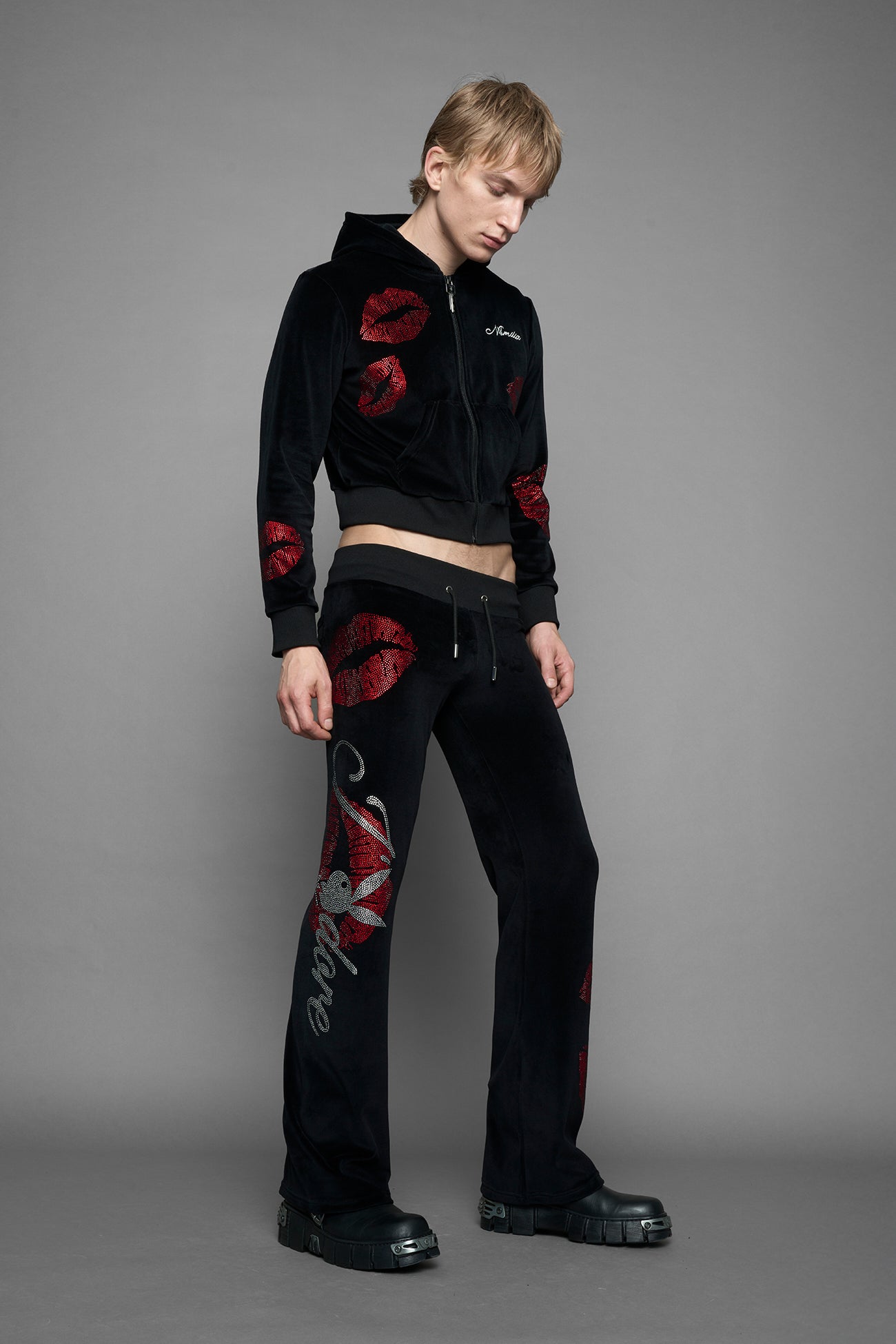 A female model wearing a black velvet zip-up hoodie with red sequined lip-print patches on the chest and sleeve, featuring white 'Namilia' embroidery on the left chest, photographed against a solid gray studio background.