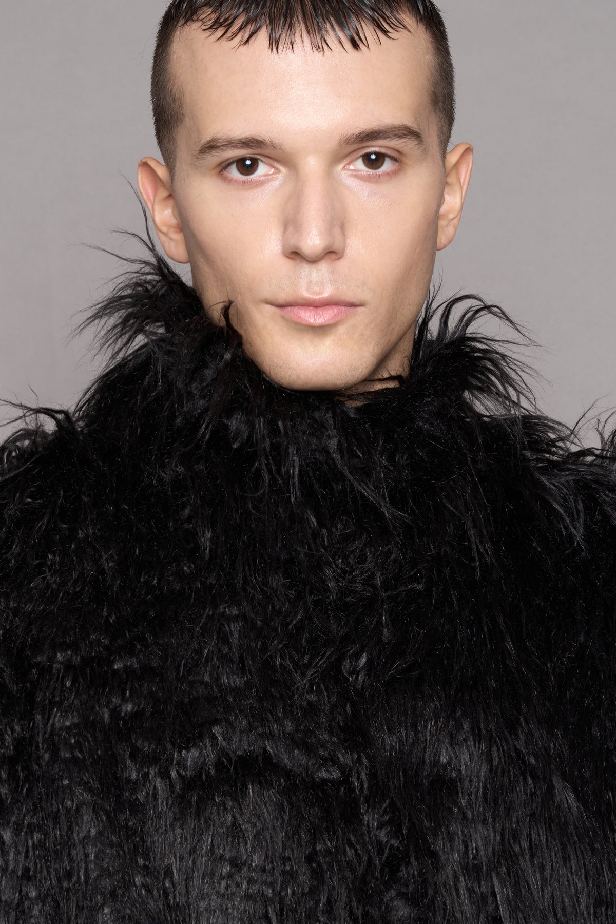 A close-up portrait of a man wearing a voluminous black faux-fur coat with a  high collar 