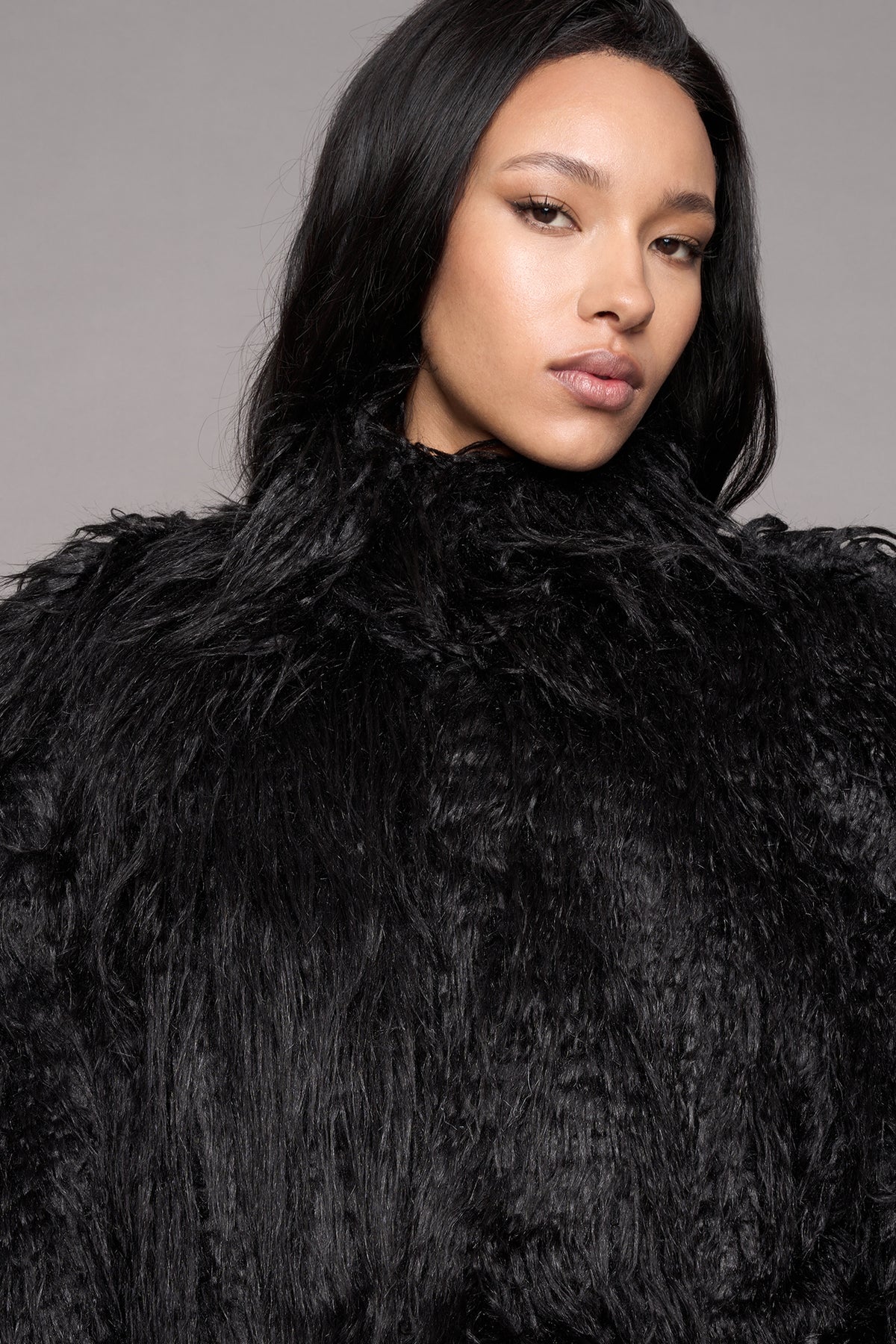 A close-up portrait of a model wearing a voluminous, shaggy black faux fur against a plain gray studio background.