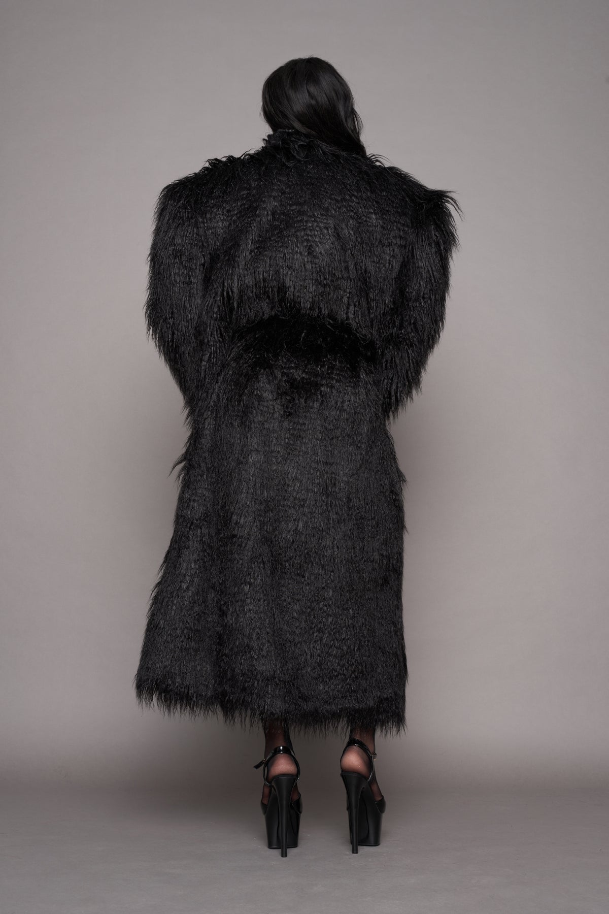 A female model viewed from behind wearing a long, voluminous black shaggy faux-fur coat with wide shoulders, paired with black high-heeled sandals, against a neutral gray studio background.