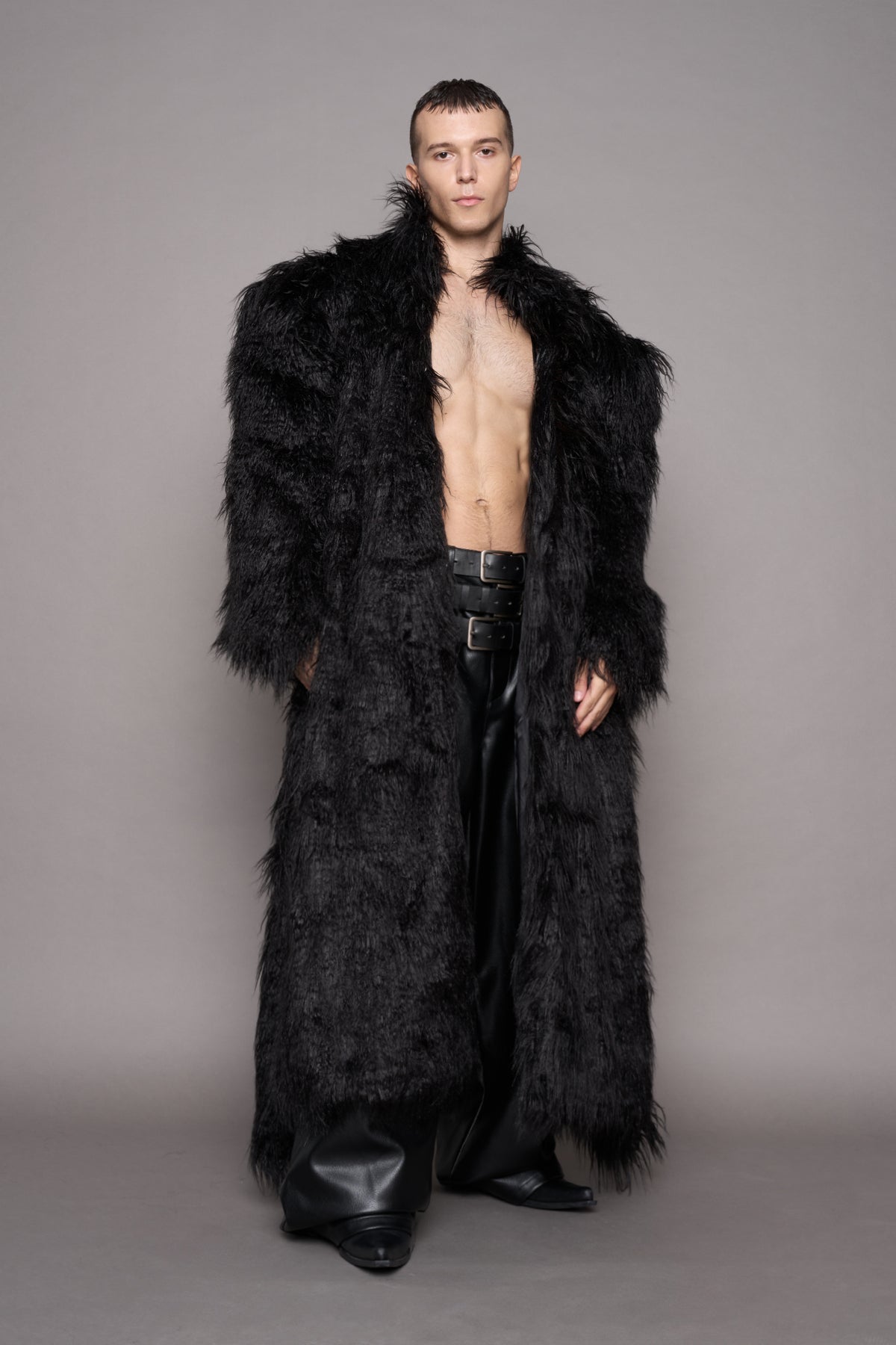 A shirtless male model wearing a long, voluminous black faux fur coat over black leather pants and boots, standing against a plain gray studio background.
