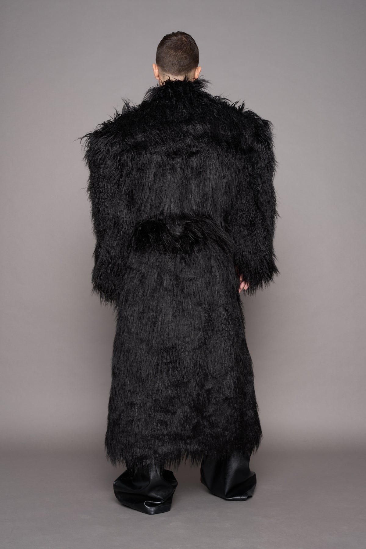 A model viewed from the back wearing a long, voluminous black shaggy faux-fur coat with wide shoulders, paired with black leather boots, against a solid gray studio background.