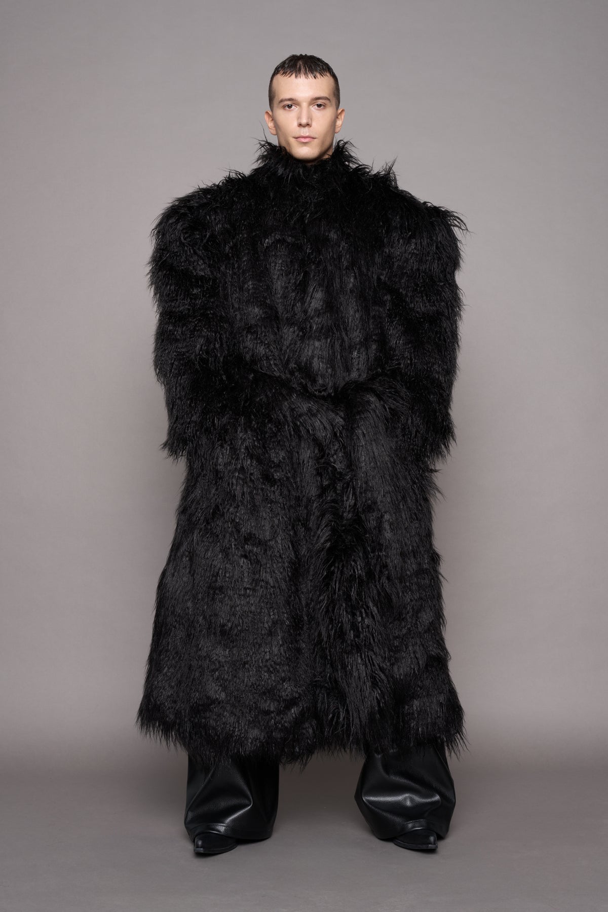 A model wearing a voluminous, oversized black faux fur coat with a high collar and a matching waist tie, paired with black leather boots, standing against a solid gray studio background.