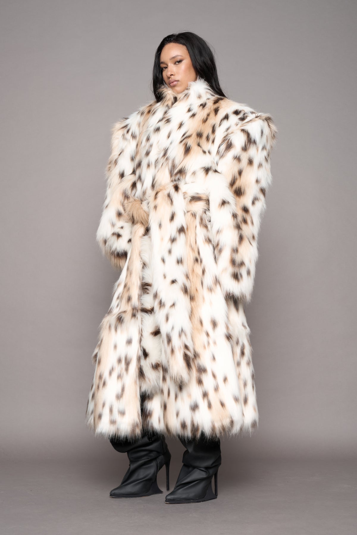 A model wearing a long, oversized faux fur coat leopard pattern, paired with black pointed-toe boots, against a solid gray studio background.