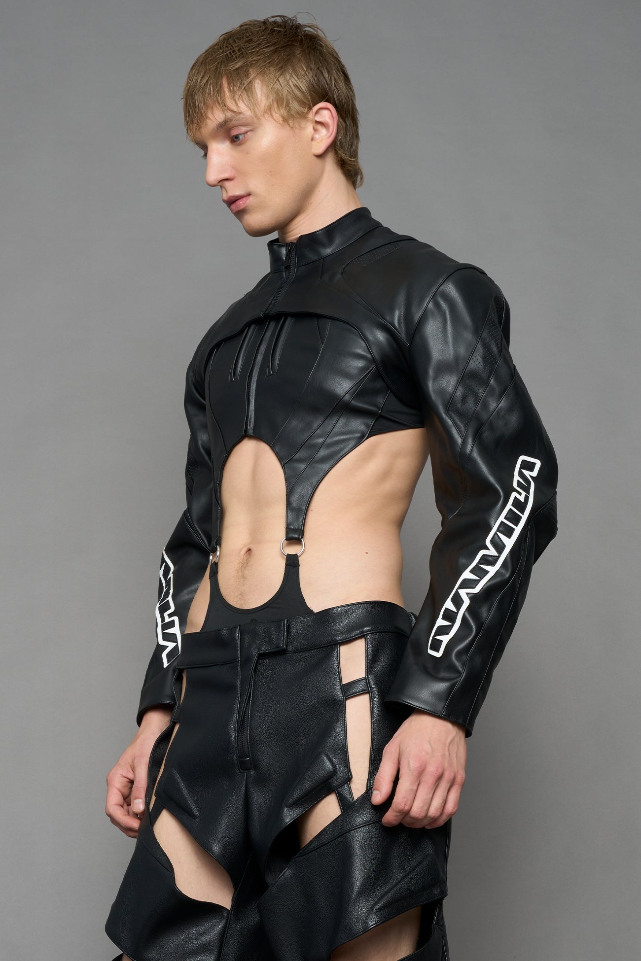 A male model wearing a black faux-leather long-sleeve top with a high collar and white graphic text on the sleeves