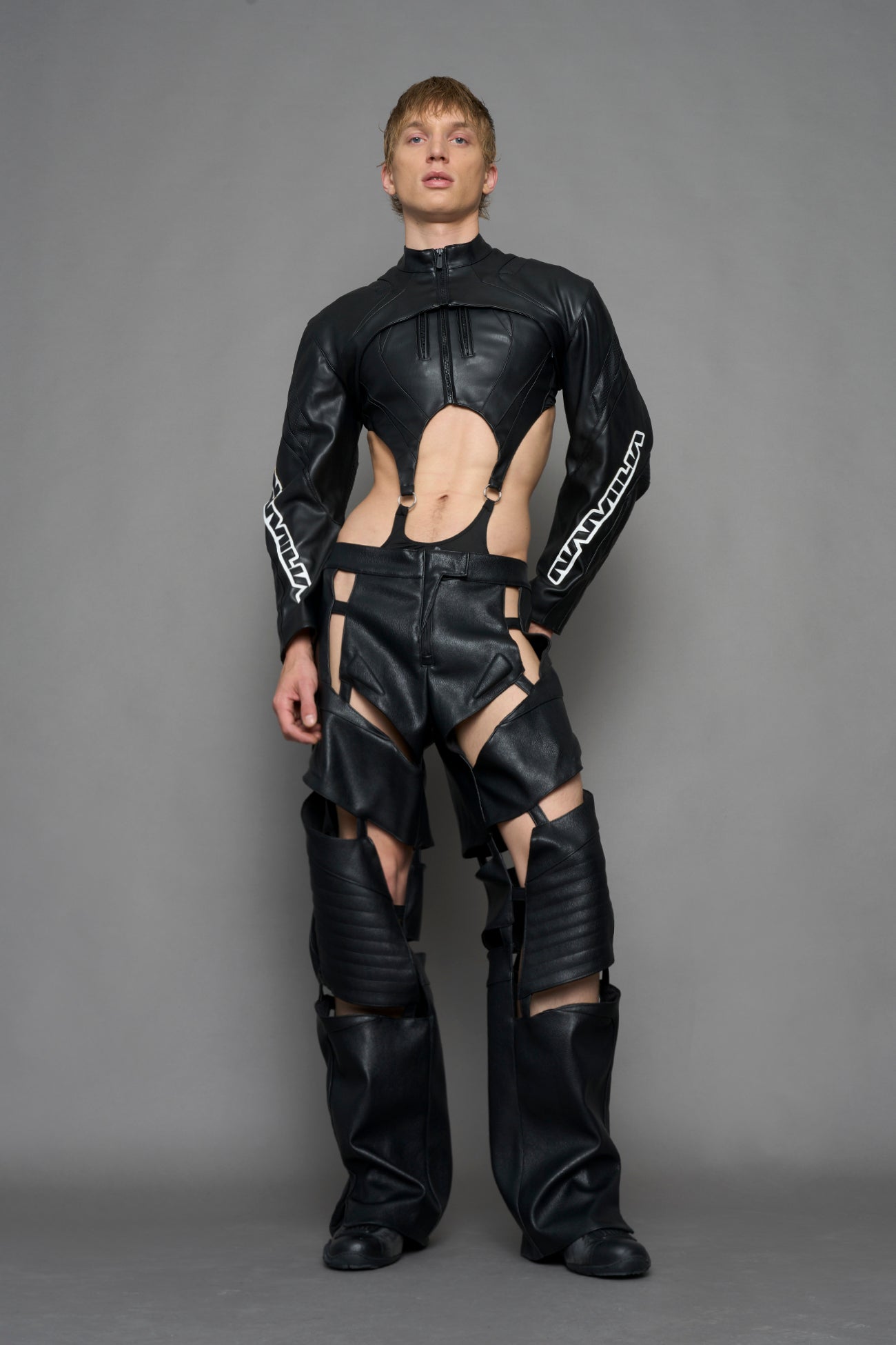 A male model wearing a black faux-leather long-sleeve top with a high collar and white graphic text on the sleeves