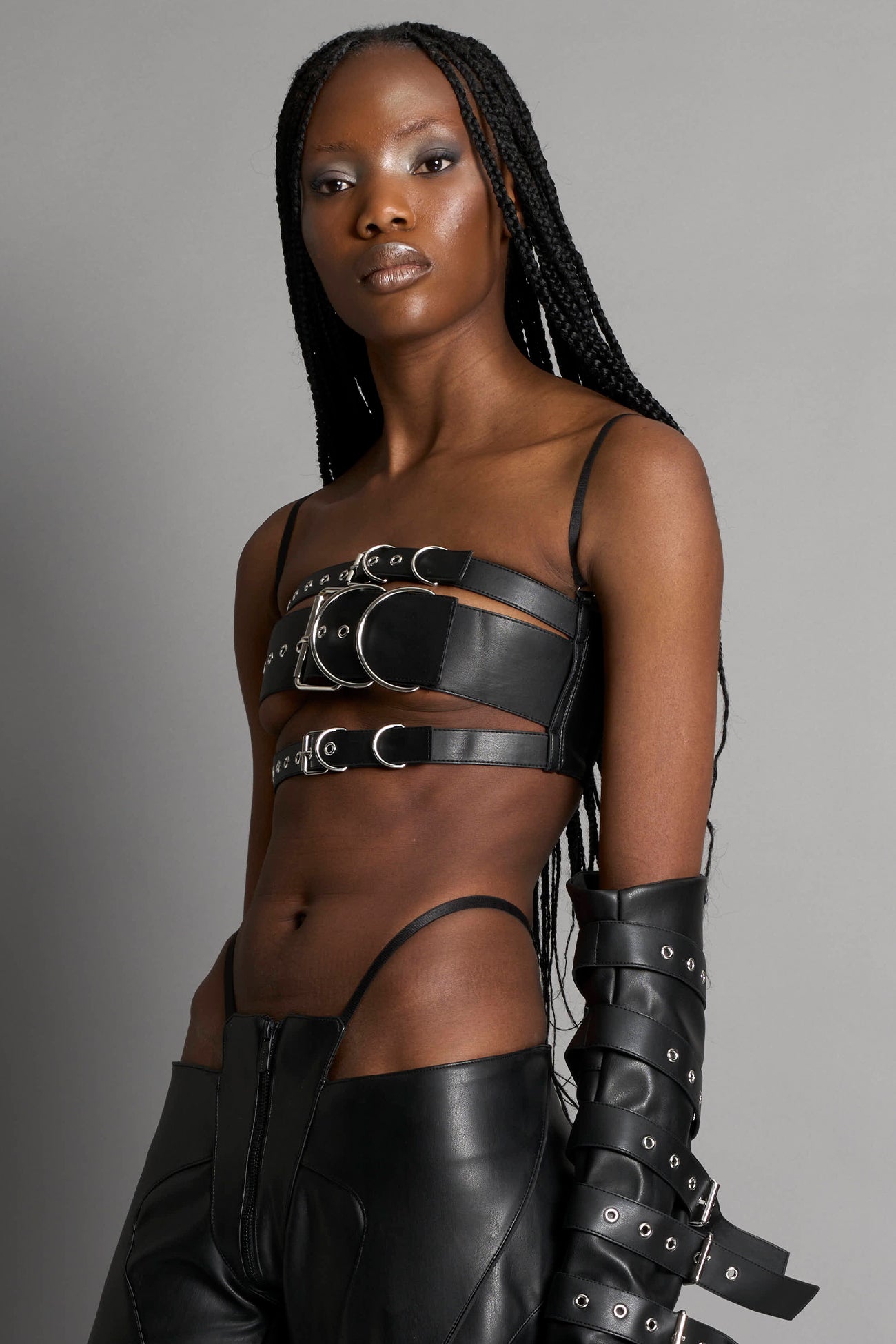 Black vegan leather belt top with three adjustable grommet straps, oversized metal buckles, and circular hardware – edgy bondage-inspired crop top for festival or clubwear.

