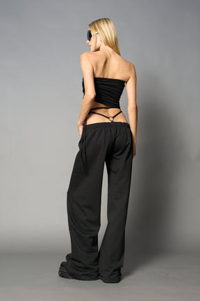 Model wearing low-rise black wide-leg pants featuring a V-shaped waistband and detachable thong strap detail.