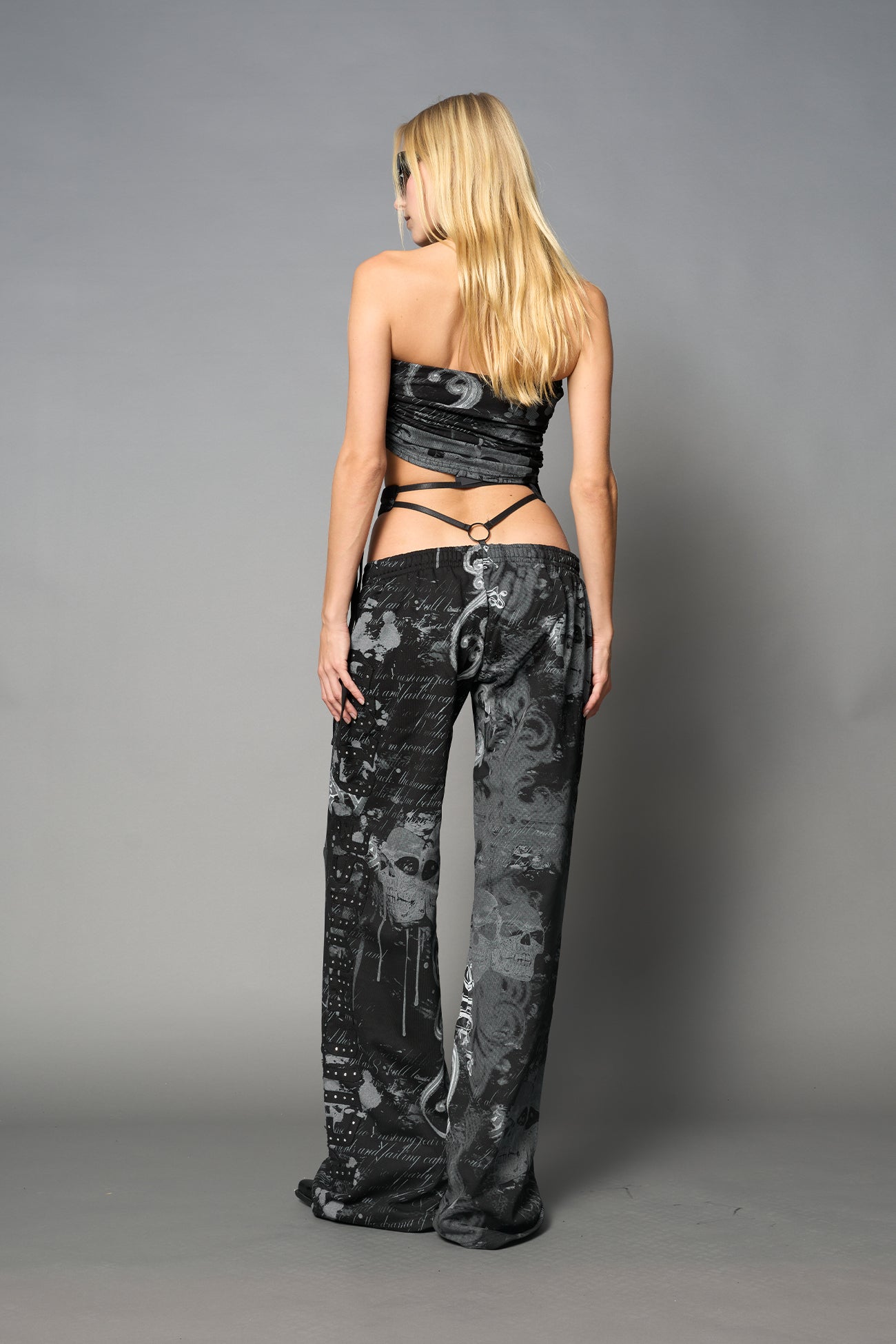 Model wearing low-rise black print wide-leg pants featuring a V-shaped waistband and detachable thong strap detail.