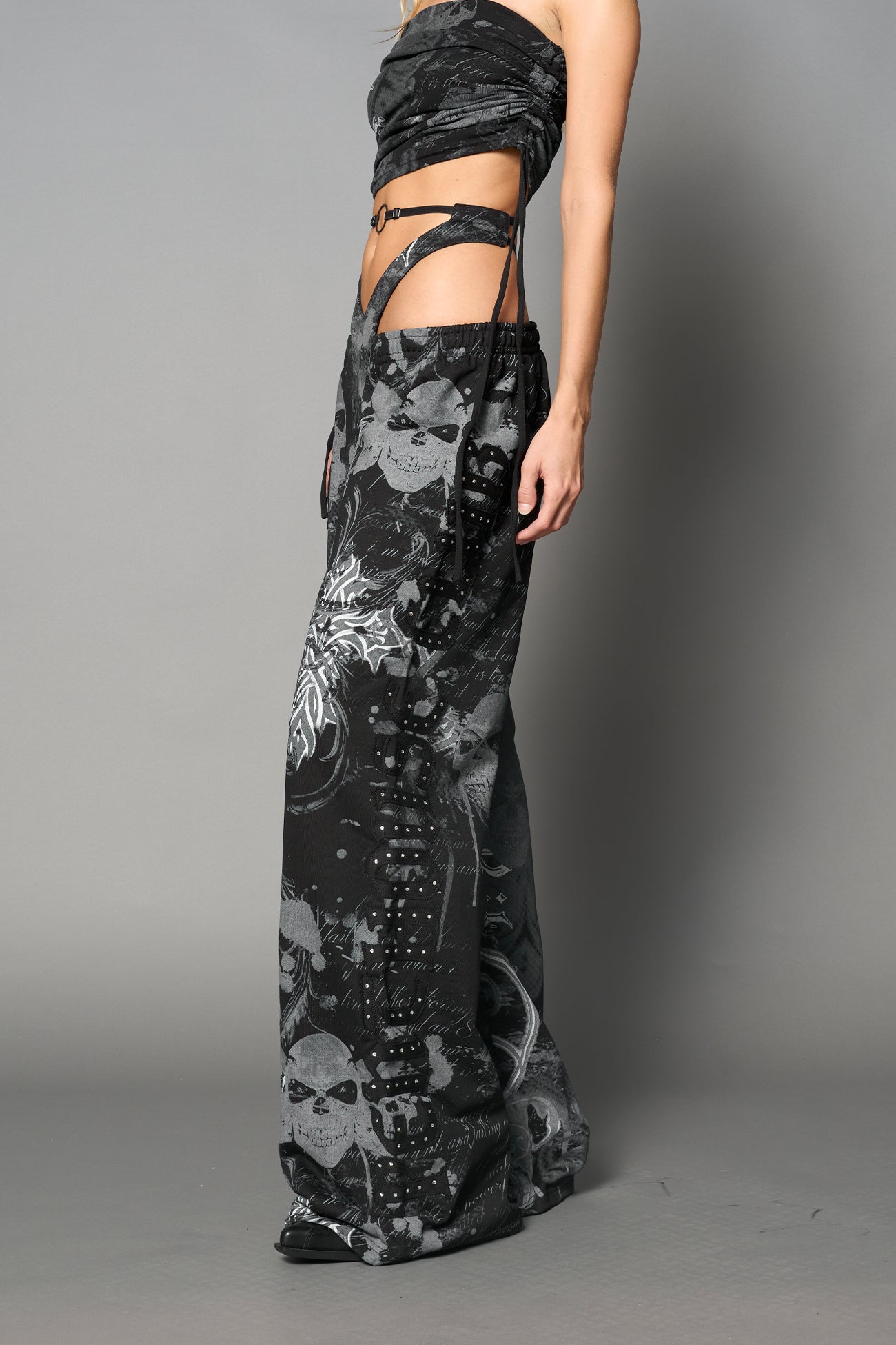 Model wearing low-rise black print wide-leg pants featuring a V-shaped waistband and detachable thong strap detail.