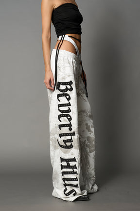 Model wearing low-rise white print wide-leg pants featuring a V-shaped waistband and detachable thong strap detail.