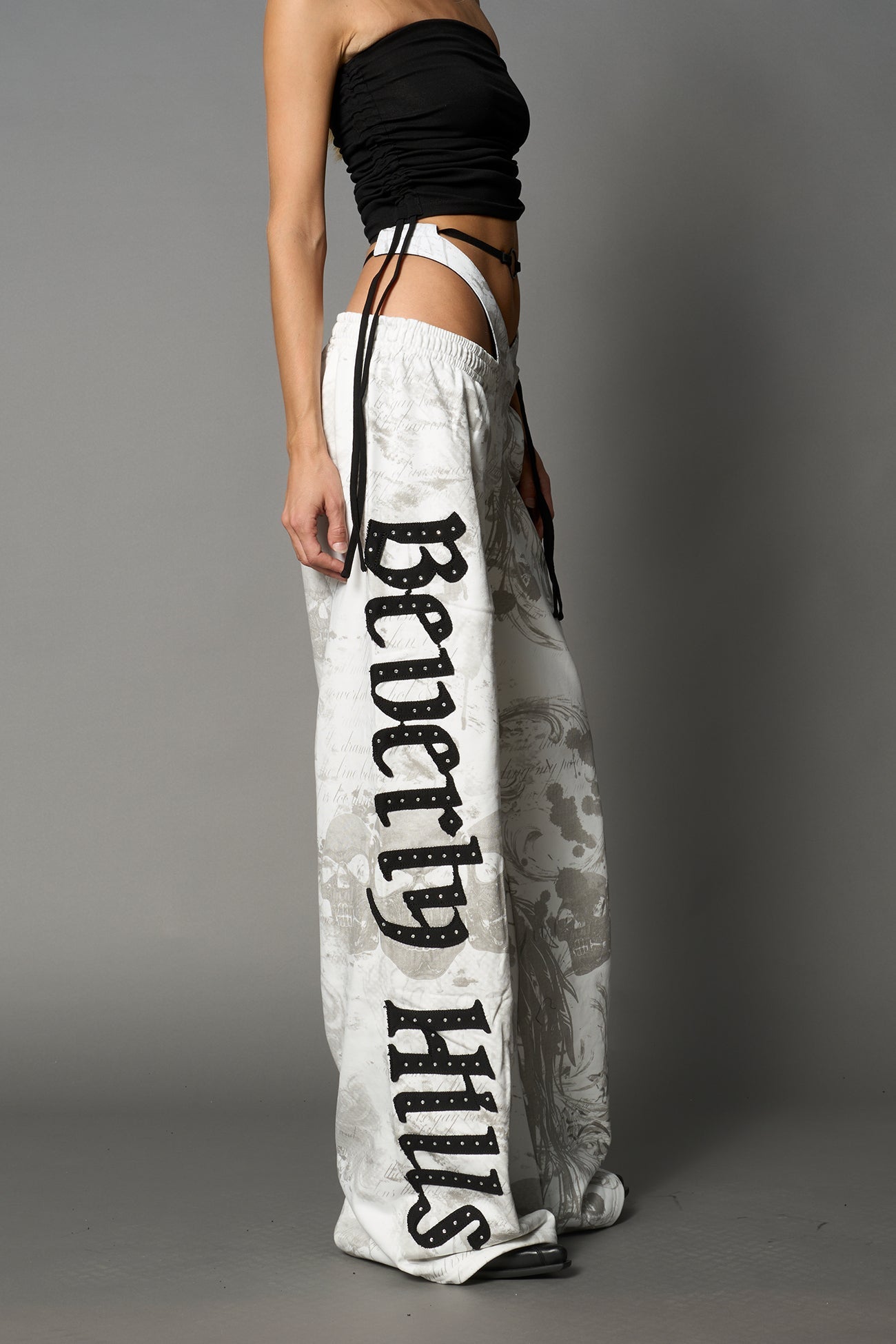 Model wearing low-rise white print wide-leg pants featuring a V-shaped waistband and detachable thong strap detail.