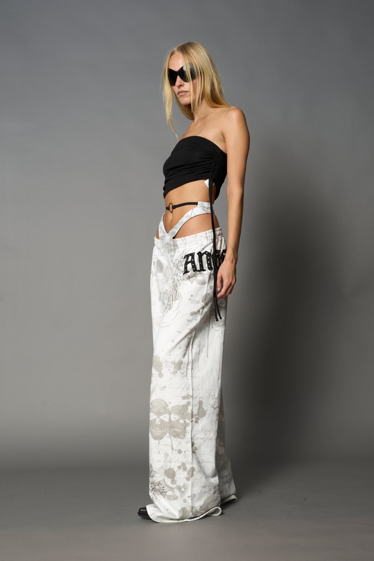 Model wearing low-rise white print wide-leg pants featuring a V-shaped waistband and detachable thong strap detail.