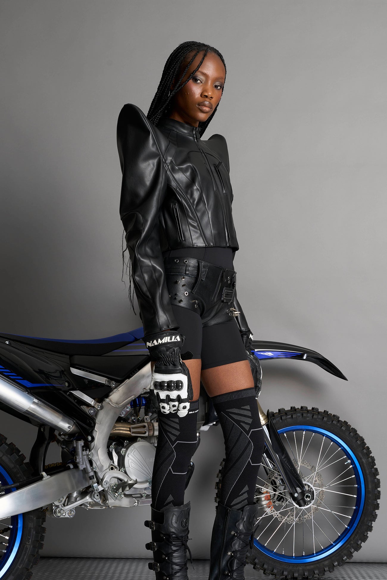 Model wears the black Namilia Spike Moto Jacket with wire shoulder pads, branded zipper, band collar, zippered breast and waist pockets, motocross-style lines, and black logo rubber tag on the back.