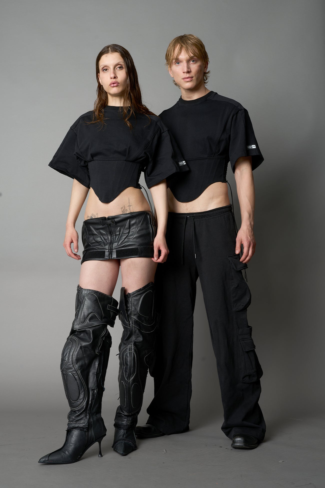 Front view of model in black Namilia Corset-T shirt with sculpted corset-style waist, curved hemline, and oversized sleeves, styled with leather mini skirt and moto boots.