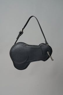 Black vegan leather shoulder bag in the shape of a phallus, featuring a silver piercing detail and an adjustable strap, displayed against a neutral grey background.