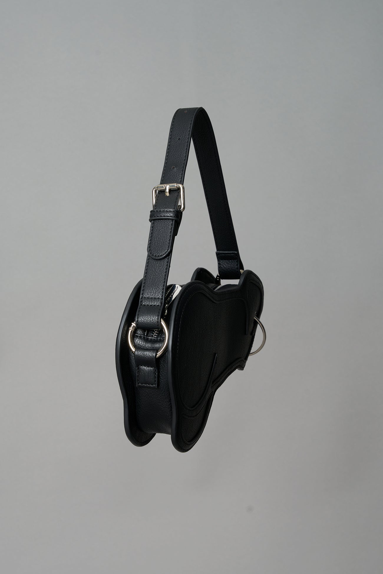 Black vegan leather shoulder bag, featuring a silver piercing detail and an adjustable strap, displayed against a neutral grey background."