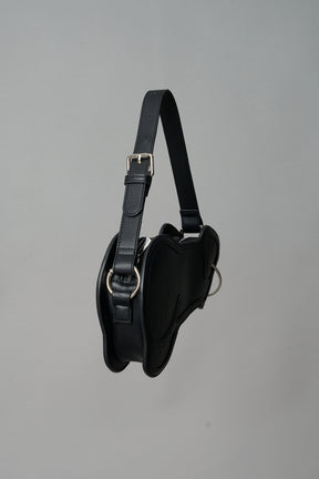 Black vegan leather shoulder bag, featuring a silver piercing detail and an adjustable strap, displayed against a neutral grey background."